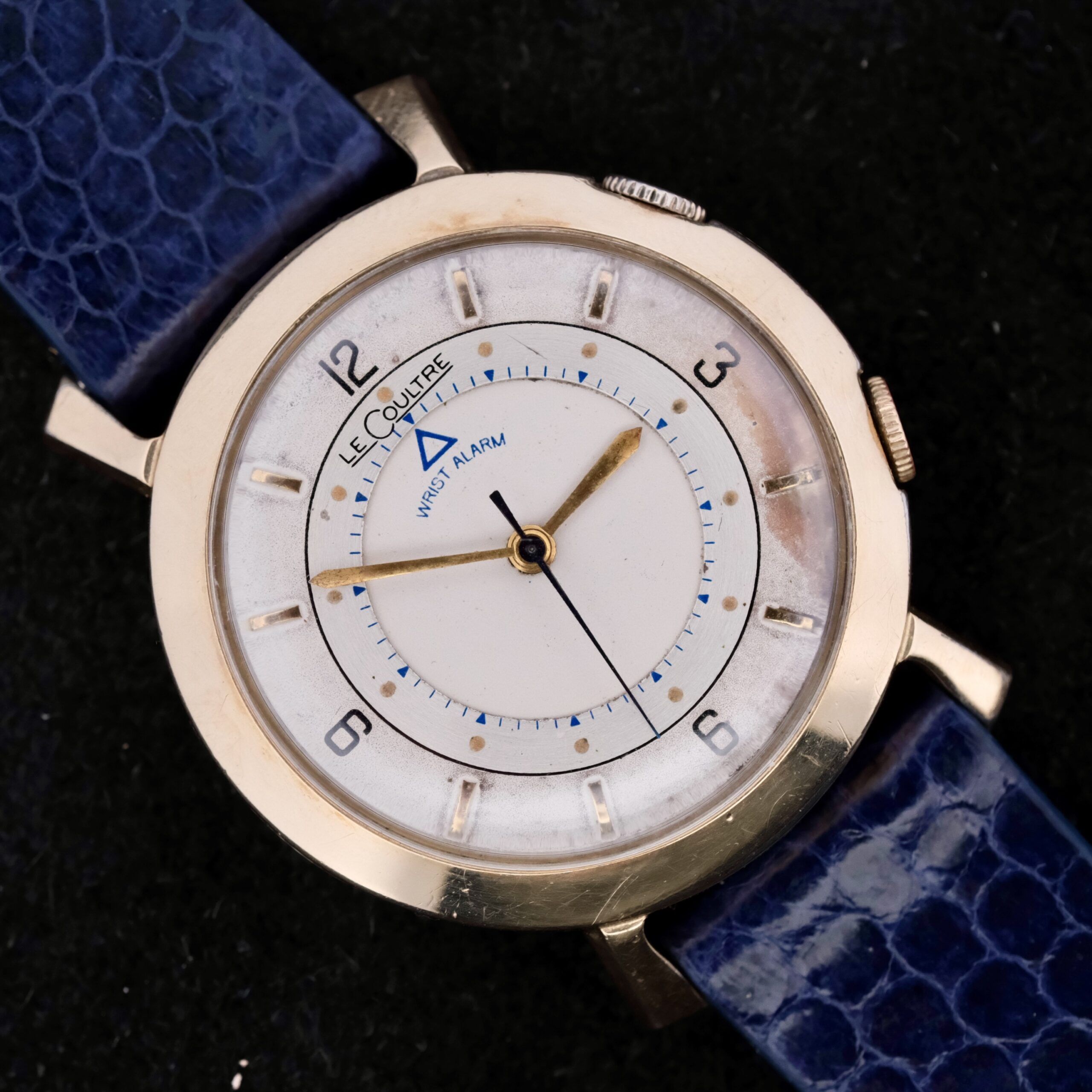 The vintage Lecoultre Wrist Alarm watch has a gold-tone case, white dial, numeral hour markers, and a blue leather strap. It features two crowns for its wrist alarm function and displays LeCoultre at the top.