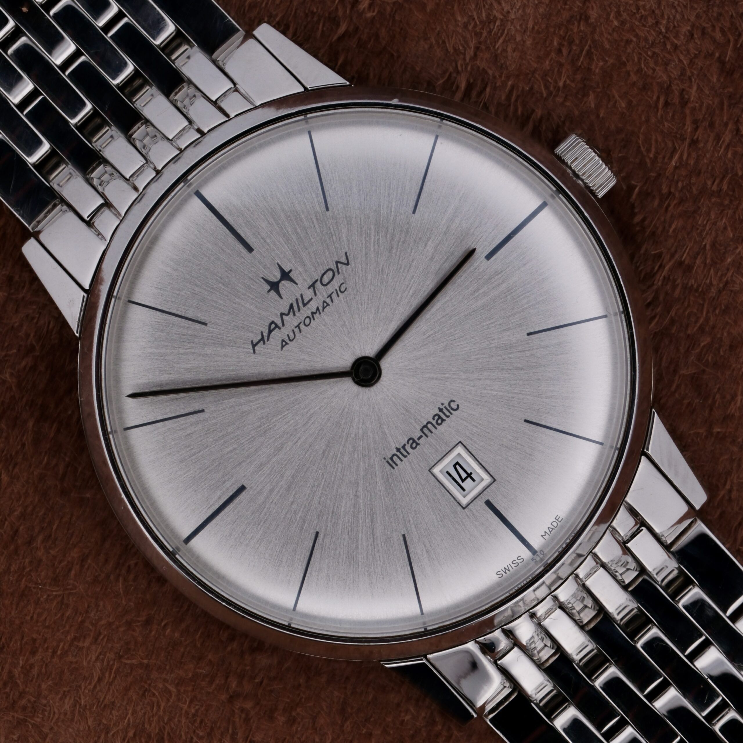 Close-up of a silver Hamilton Intramatic Date Stainless Steel automatic wristwatch with a stainless steel bracelet. The face shows black hour markers, two hands, and a date display reading 14th, set against a brown background.