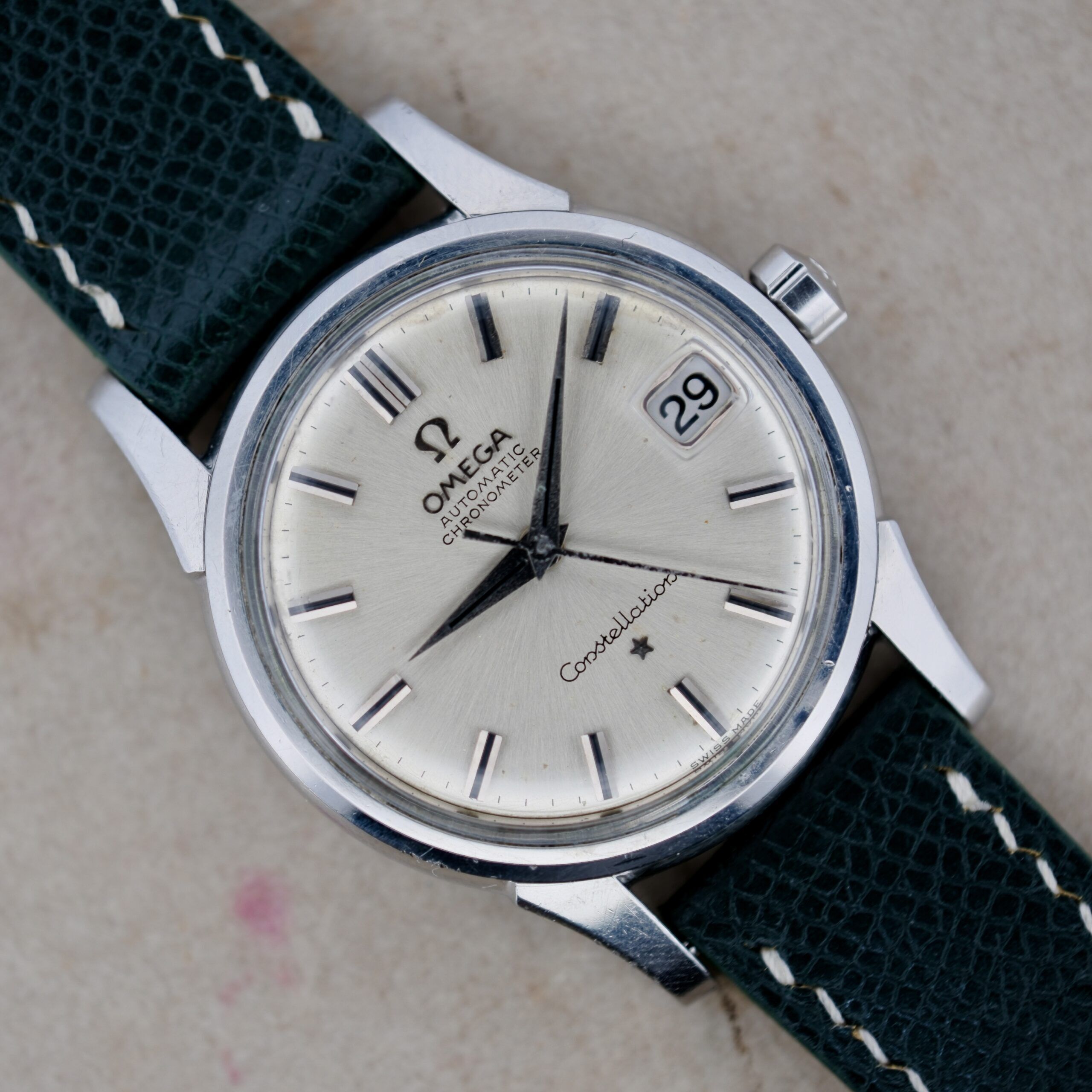 The Omega Constellation Chronometer 14939-1 Flat Dial has a silver round face with black hour markers, hands, and a date display. It features a textured leather strap with white stitching, while the face reads Omega Automatic Chronometer Constellation, exuding timeless elegance.