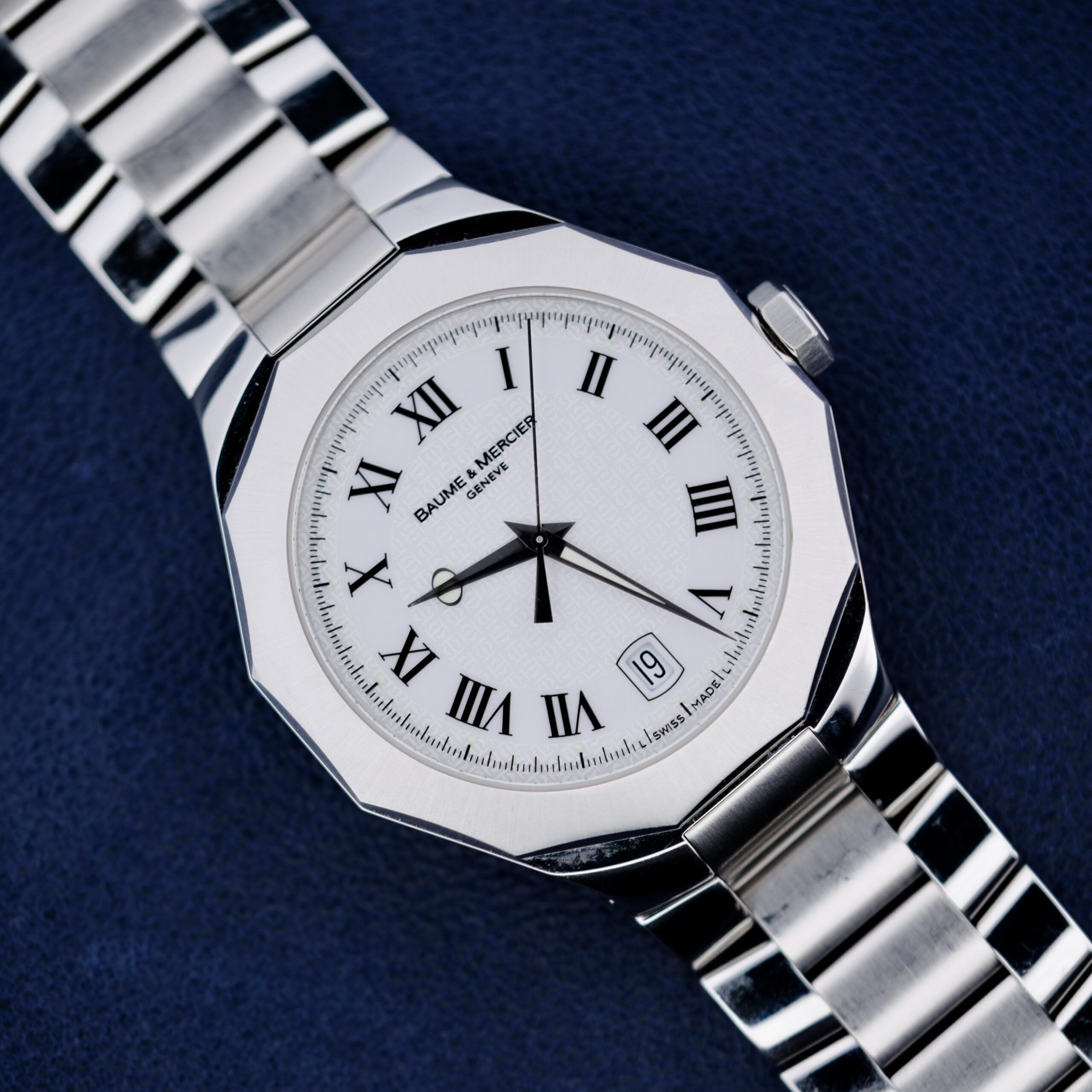 The Baume Mercier Riviera 65580 is a silver wristwatch featuring a round white face with Roman numerals and a date window. It is displayed on a dark blue textured surface, with its polished metal bracelet completing the elegant look.