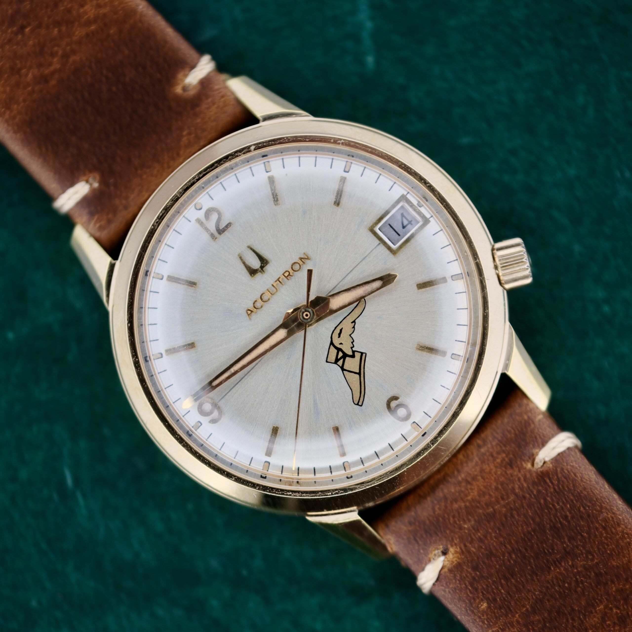 A Bulova Accutron 218 Goodyear Dial watch with a brown leather strap rests elegantly on a green surface. The silver face features gold hour markers, hands, a small date window at 3 oclock, and an intricate illustration near 6 oclock.
