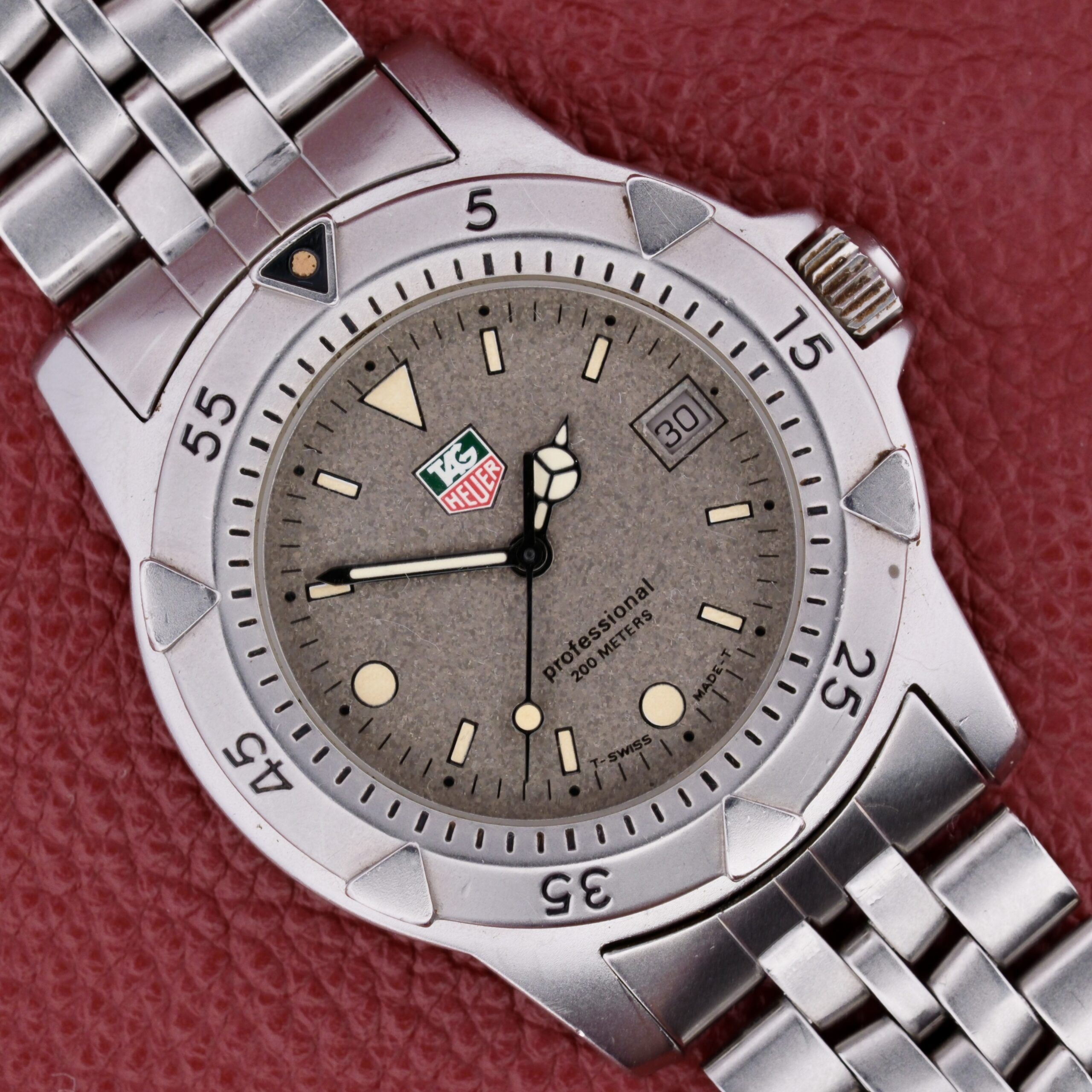 A Tag Heuer 1500 Series wristwatch with a silver metal bracelet sits on a red textured background. It features a round face, gray dial, white hour markers, and a date display at 3 oclock, blending sporty design with elegance.