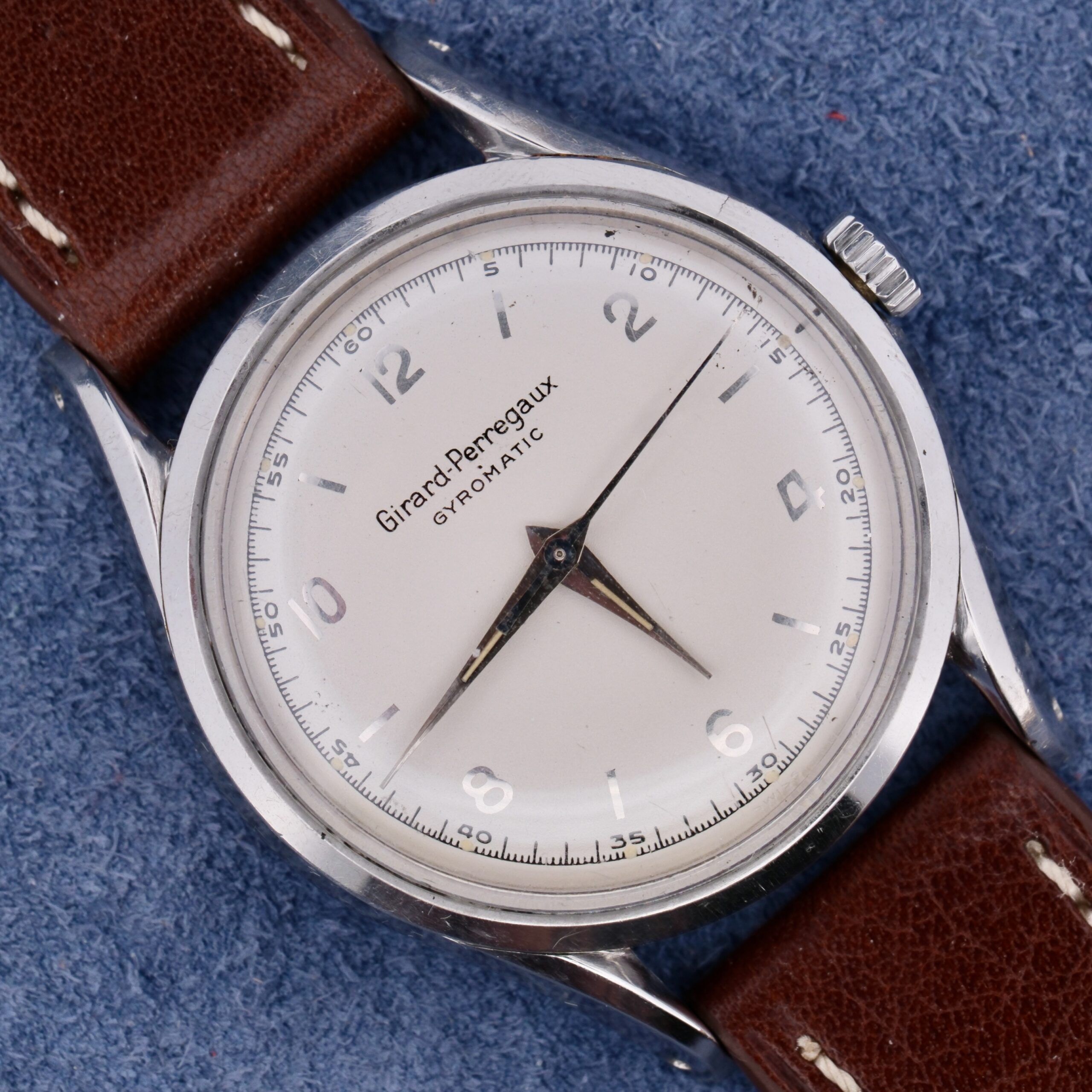 The vintage Girard Perregaux Gyromatic 6169 wristwatch showcases a white dial, silver hour markers, and a brown leather strap, elegantly set against a blue textured background.