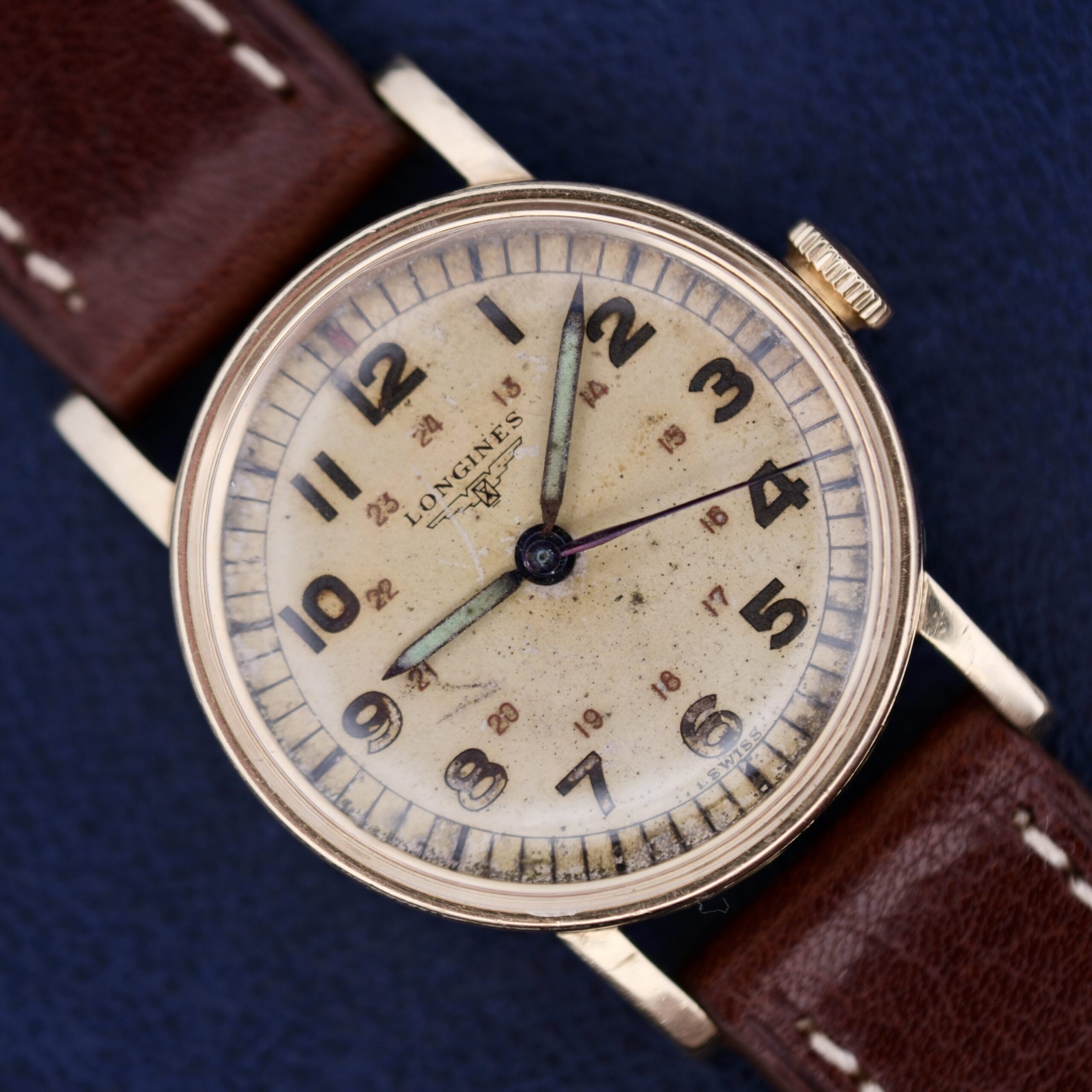 A classic Longines Military 24 Hour Dial 12L wristwatch features a round-aged white dial with black numerals and blue hands. It has a brown leather strap and displays visible wear, enhancing its vintage charm.