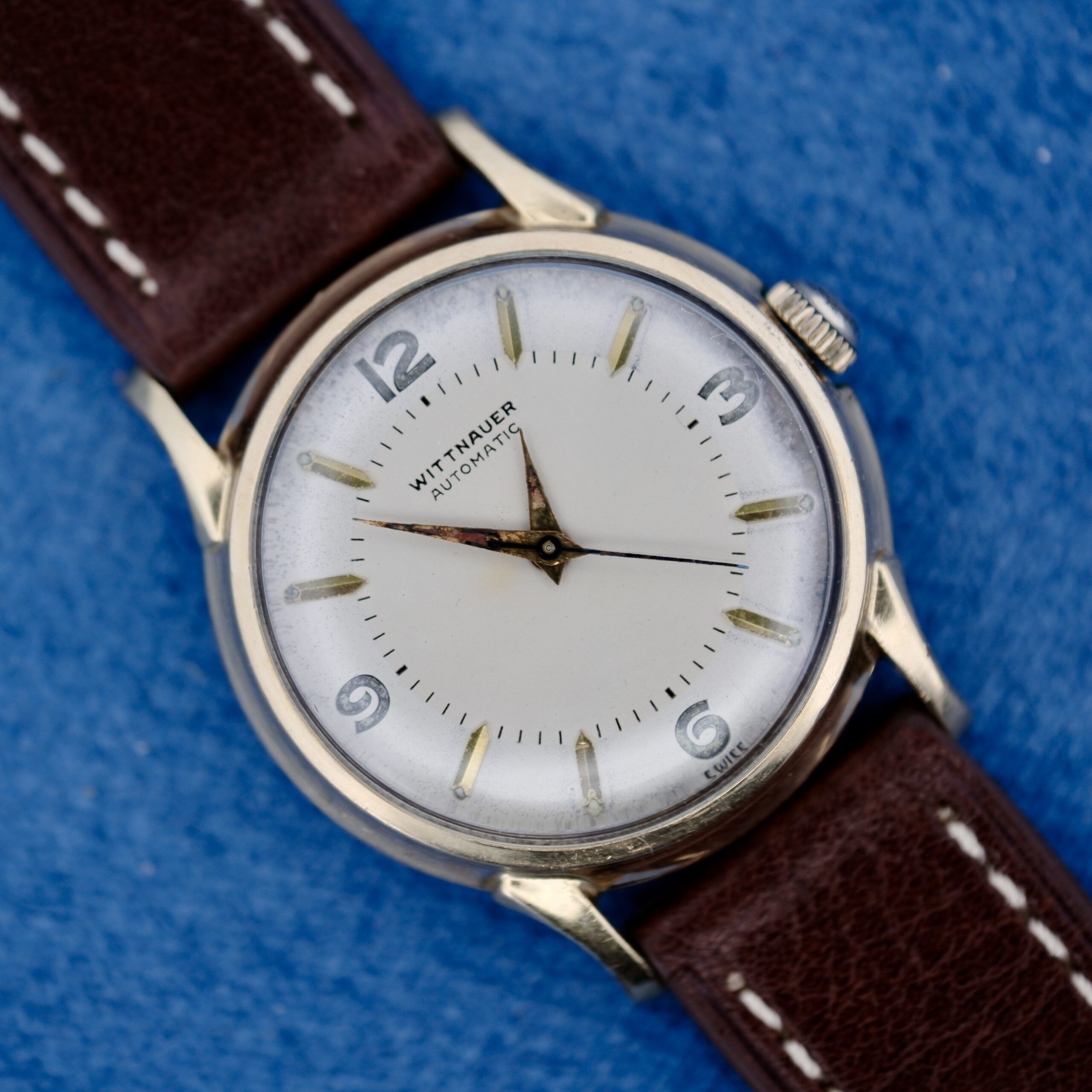 A 1950s Wittnauer Automatic Teardrop Lugs wristwatch with a white dial, silver case and hour markers, and brown leather strap is displayed on a blue textured surface, showing the time at approximately 9:14:04.