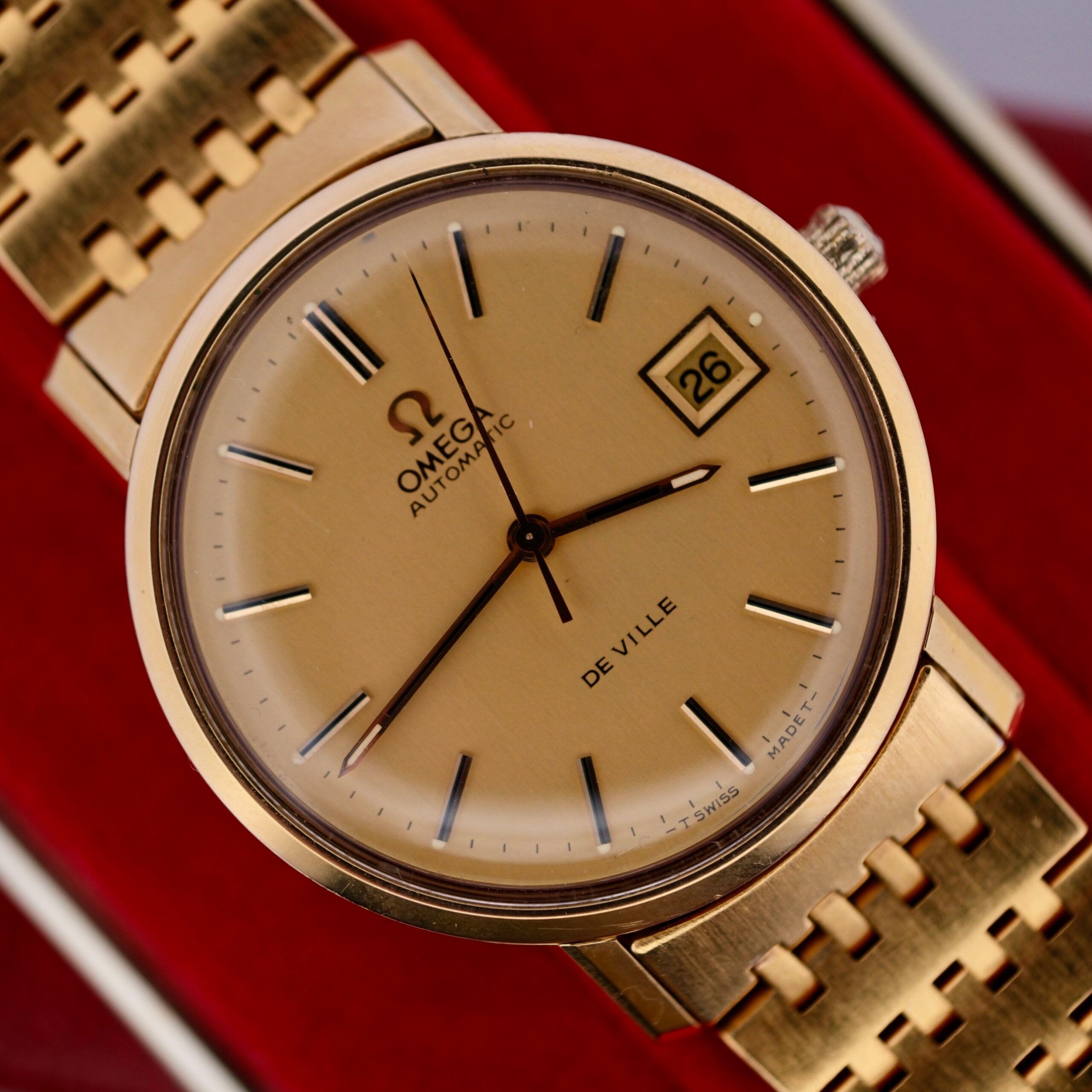 A close-up of the Omega Deville Automatic Gold Tone Date Full Set wristwatch highlights its matching bracelet, black hour markers, a date window showing 26 at 3 oclock, and the iconic Omega logo with AUTOMATIC and DE VILLE.