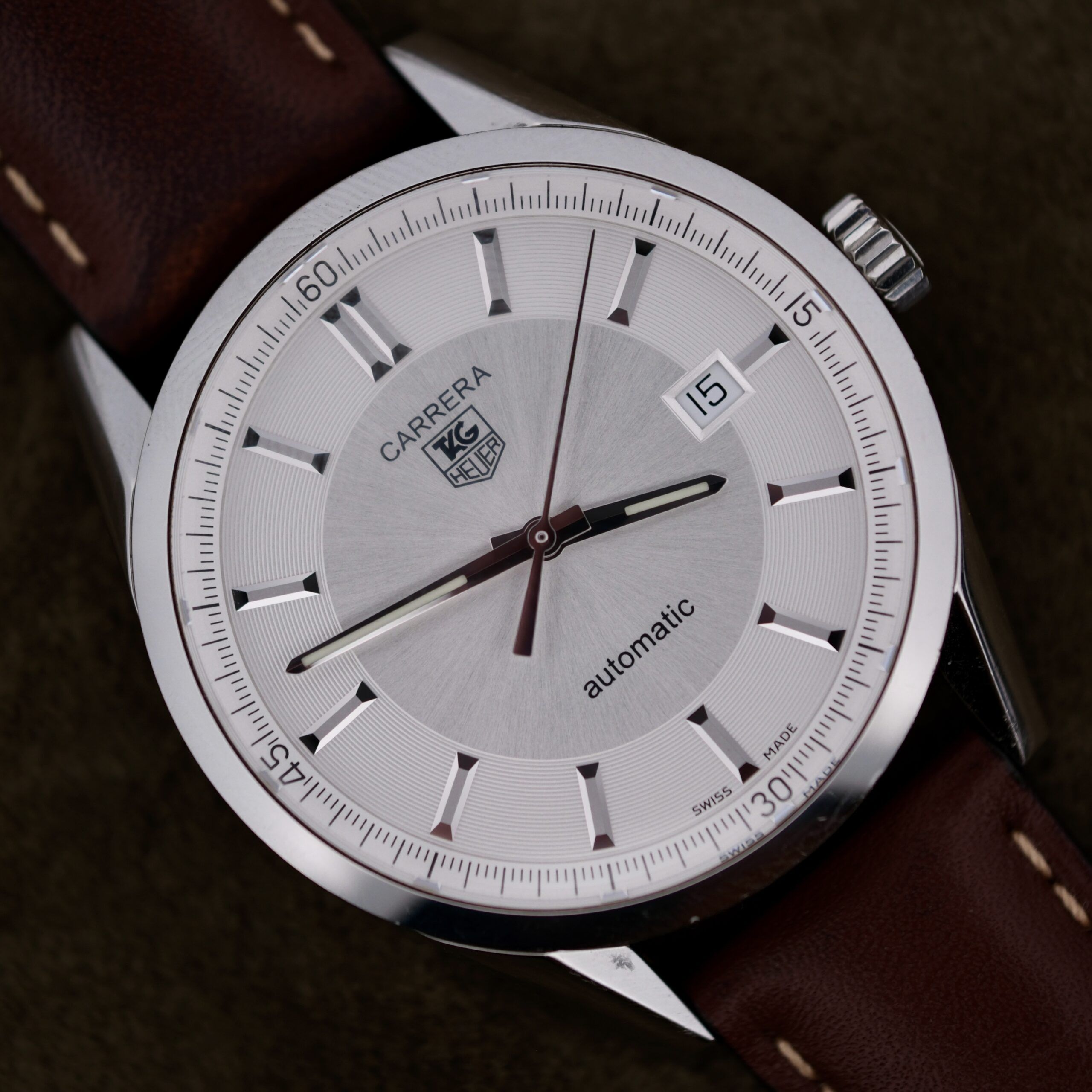 The TAG Heuer Carrera Caliber 5 Automatic Wv211A.Fc6203 features a silver dial with silver hands and hour markers, a date display at 3 oclock, the word automatic on the dial, and a brown leather strap.