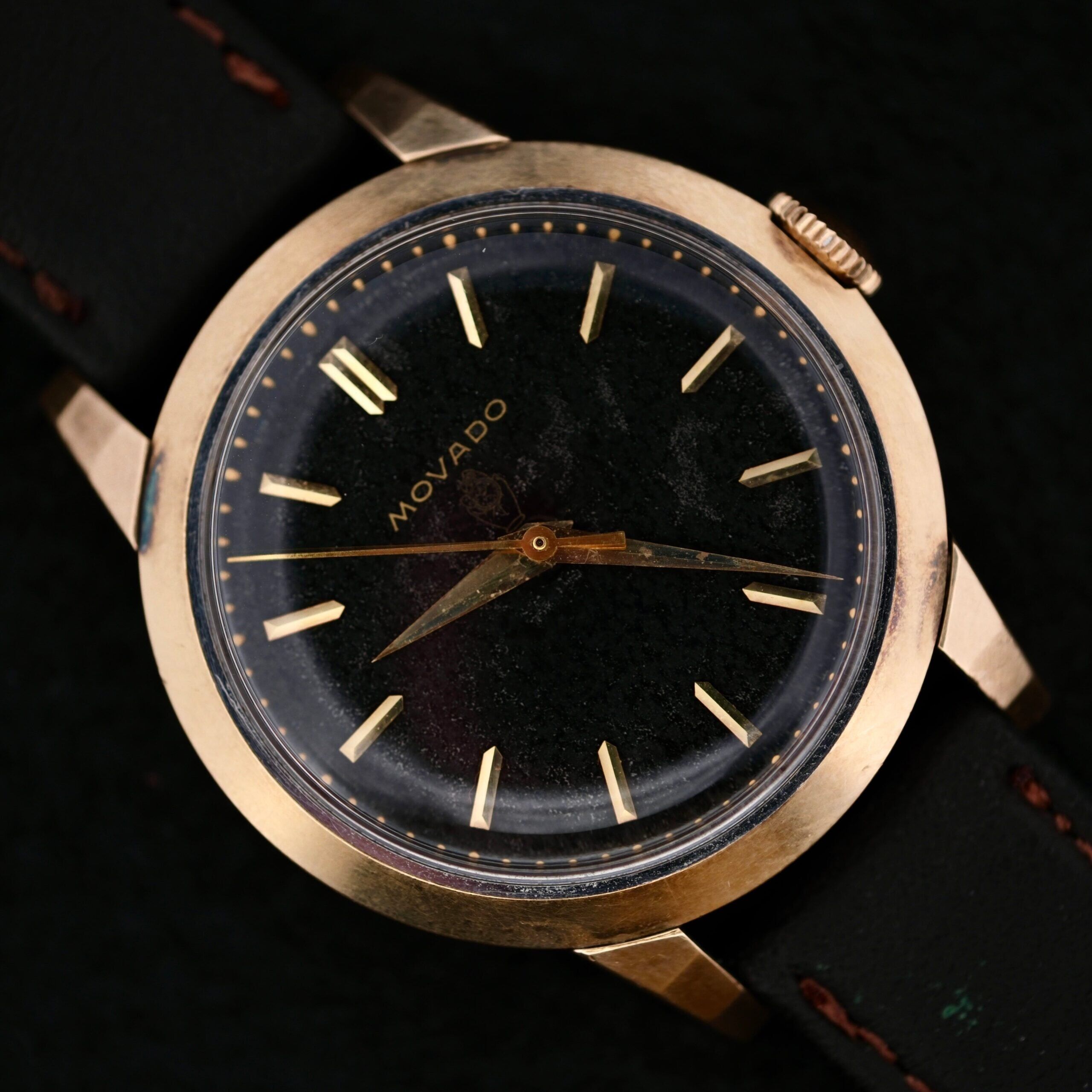A vintage Movado Ref. 18240 Francois Borgel Gold Top Hong Kong Market watch with a black face, gold case and markers, and a black leather strap. Simple design without numerals; visible wear on the glass and case enhances its classic charm.