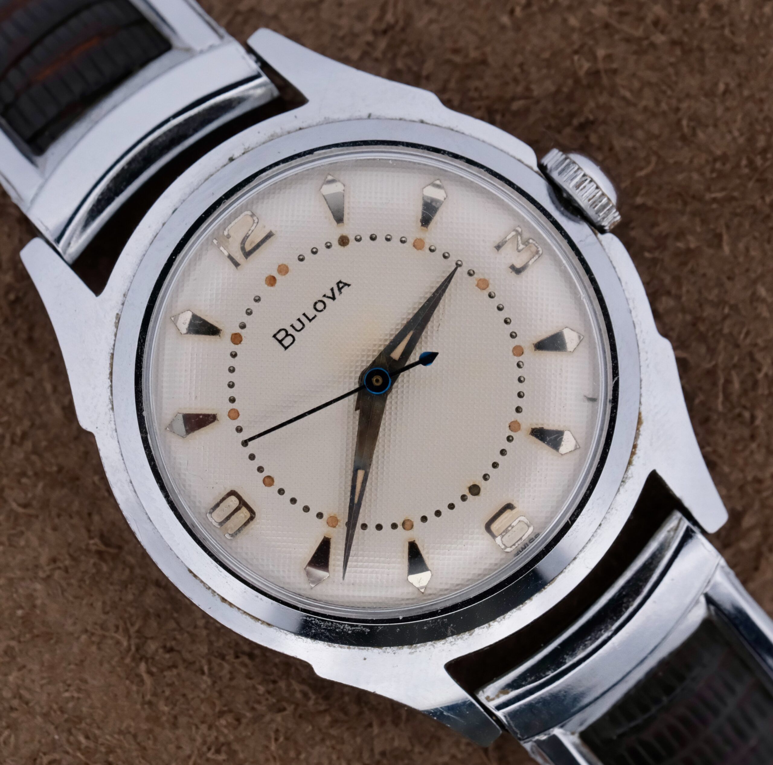 Close-up of a vintage 1956 Bulova Manual Wind Calibre 11AC wristwatch featuring a silver strap, cream-colored waffle dial, silver hour markers, numbers at 12, 3, 6, and 9 oclock, and two hands alongside a blued seconds hand on a textured brown background.