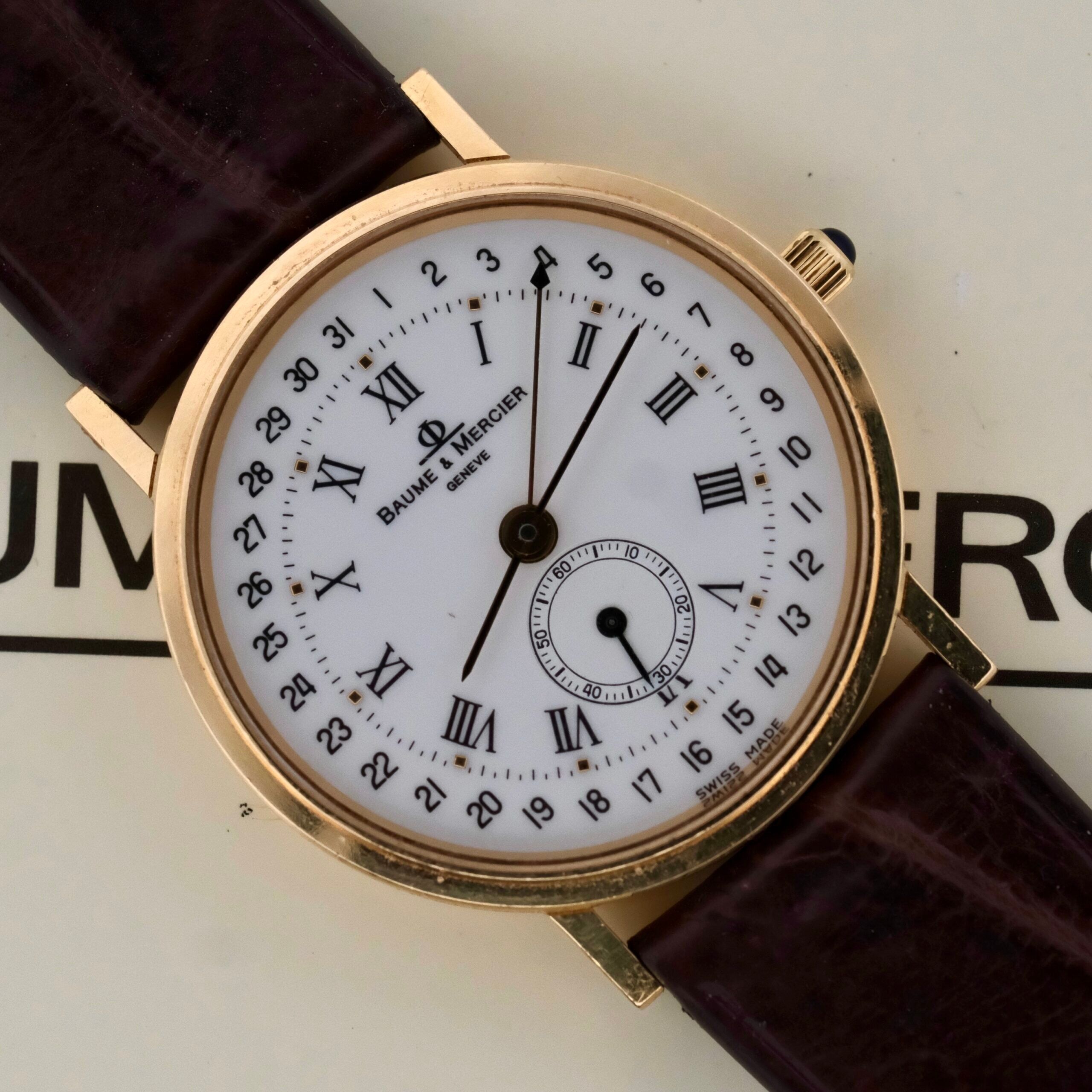 A Baume Mercier 18k Gold Calendar wristwatch (reference 15162) features a white dial with black Roman numerals, a brown leather strap, and a small seconds sub-dial above the 6 oclock position.