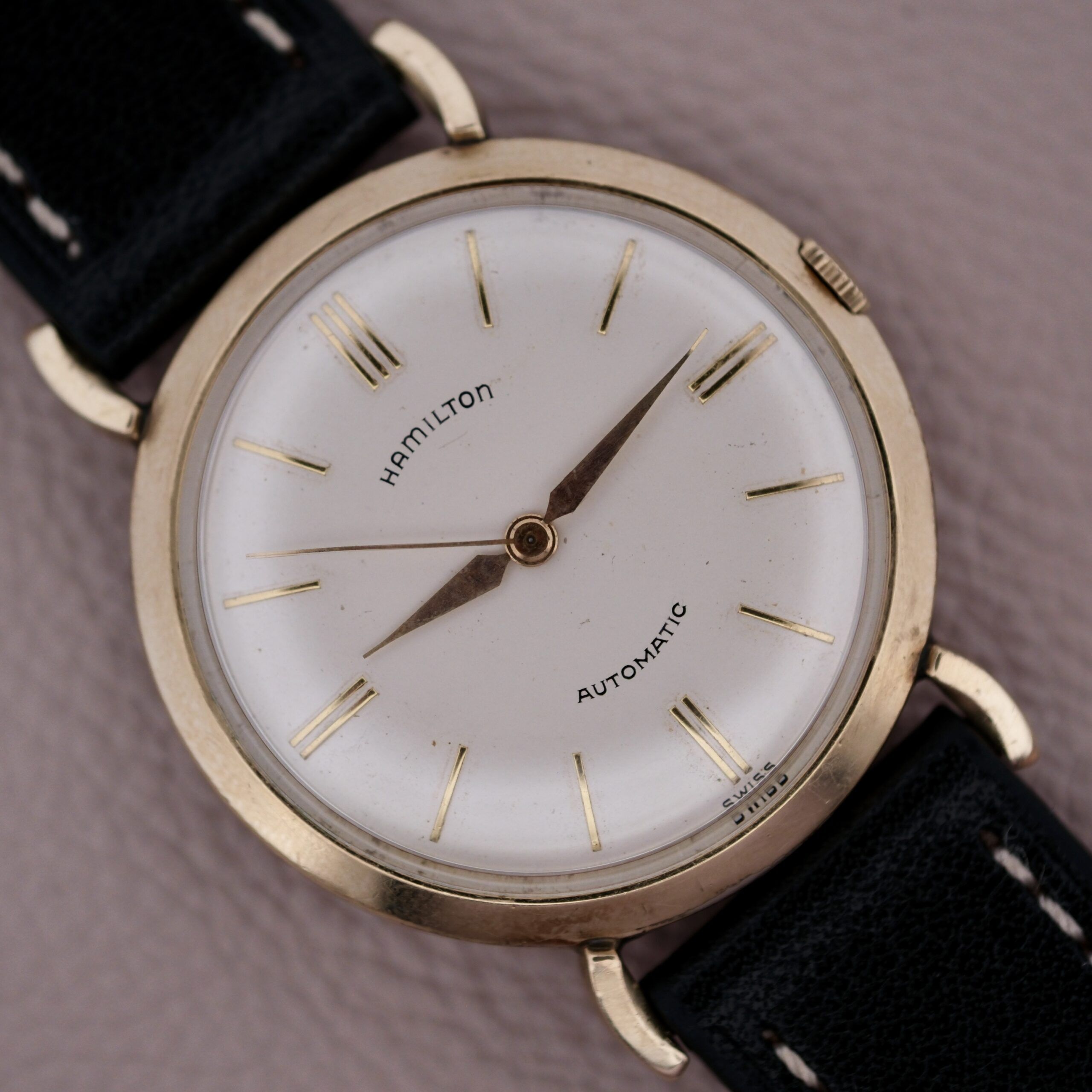 Close-up of a vintage Hamilton Kinematic Swiss Automatic Fancy Lugs watch featuring a white dial, gold-tone case, and black leather strap. Elegant details include minimalist gold hour markers and hands, with Hamilton and Automatic inscribed on the dial.