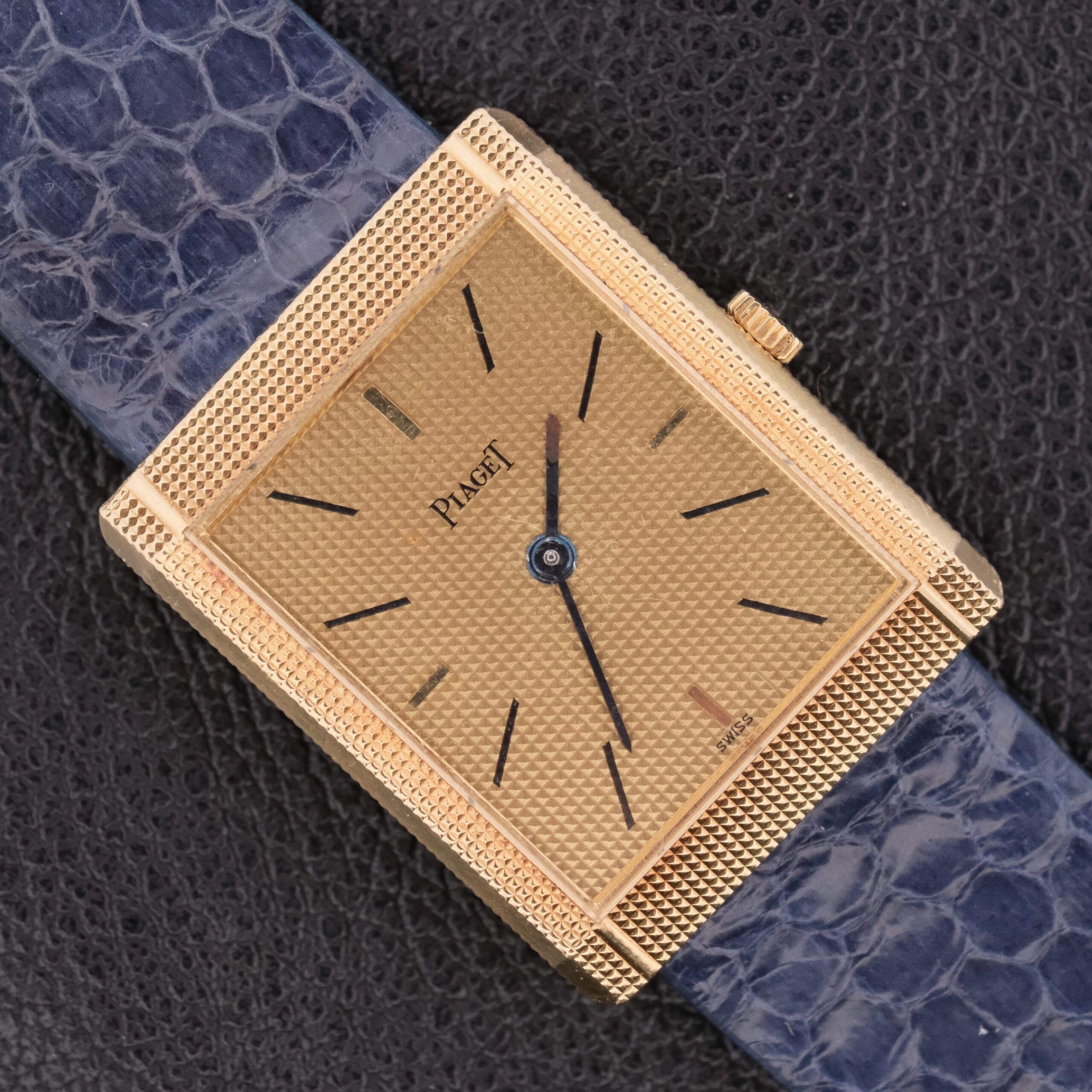 The Piaget Tank Ref 9151 18k Gold Manual Wind features a square gold watch with a textured dial, minimal black hour markers, and a blue leather strap. Its elegant design against a dark background makes it a timeless statement piece.