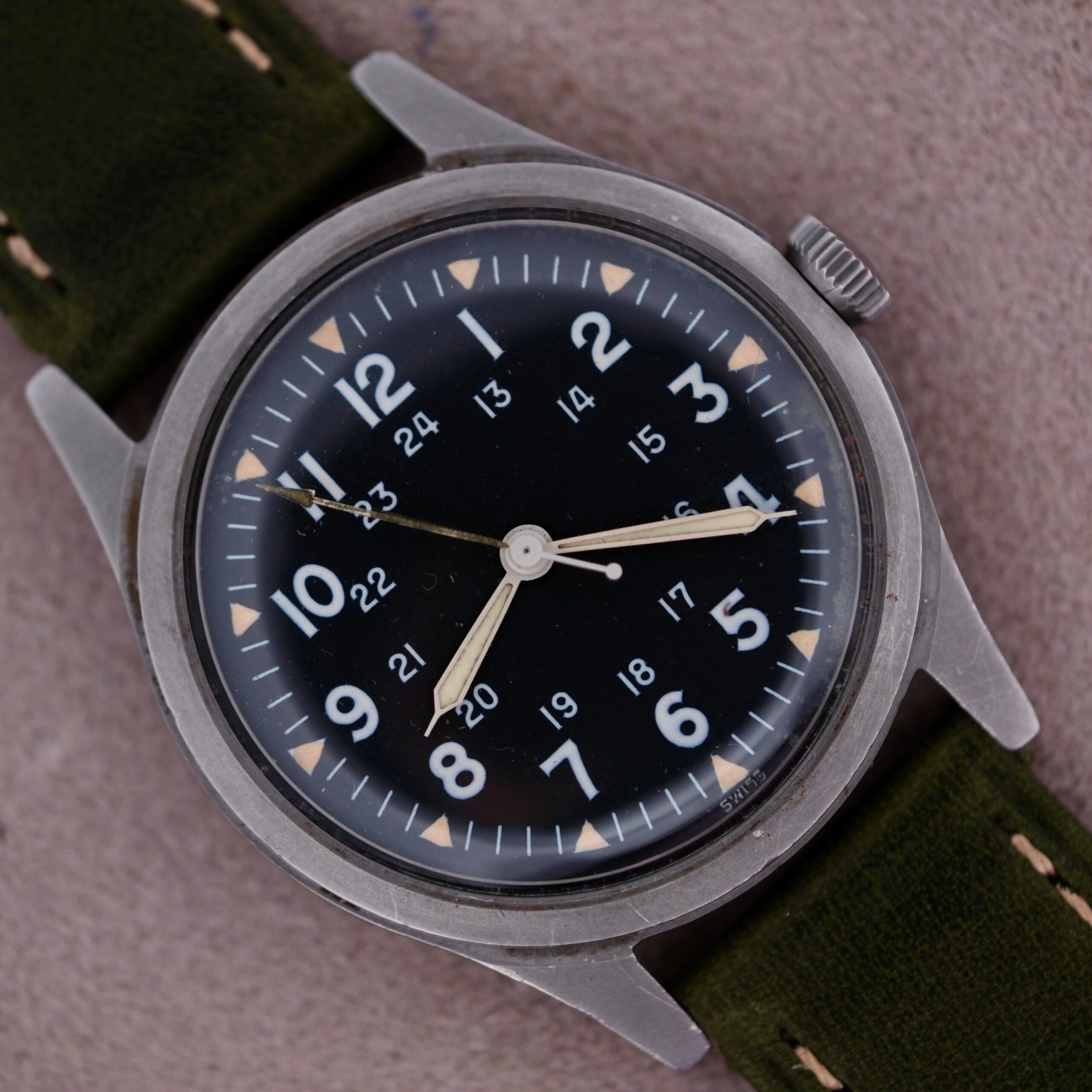 The 1964 Benrus U.S. Military Issued DTU-2A/P wristwatch features a silver metal case and green fabric strap. Its black dial displays white 12 and 24-hour markers, with luminous hands, hack second feature, and hour markers encircling the dial in a vintage design.