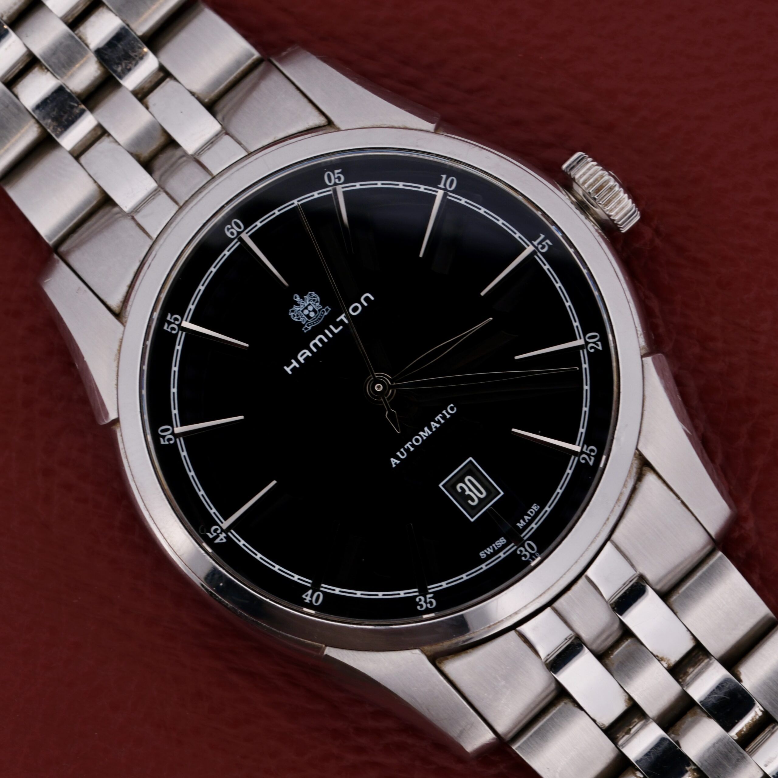 The Hamilton American Classic Spirit Of Liberty watch features a black dial, silver hour markers, and a date display at 6 oclock. It showcases an elegant stainless steel bracelet and polished bezel, complemented by a dark red leather background.