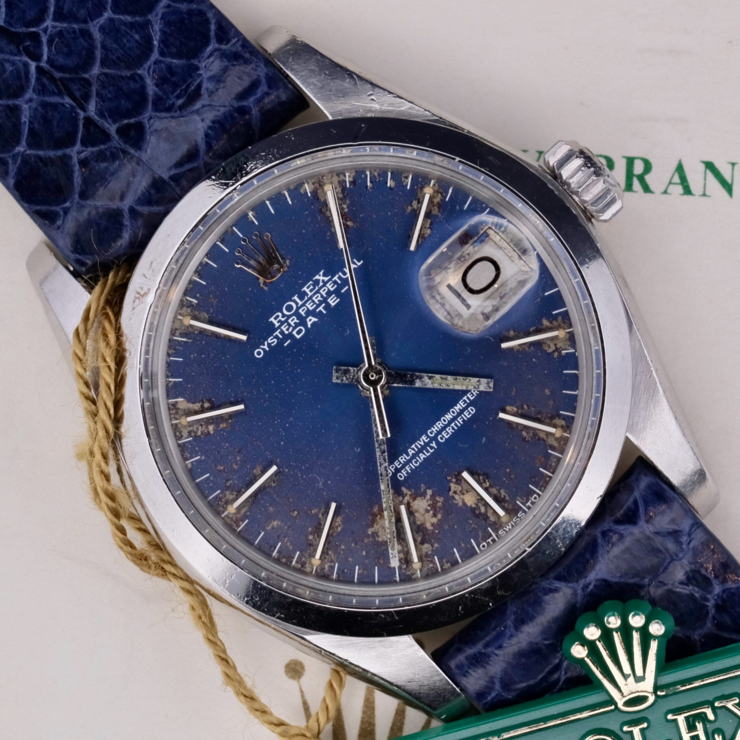 The Rolex Oyster Perpetual Date 1500 features a vintage blue, weathered dial with a date window, silver hands, and indices. It includes a blue leather strap and iconic crown logo, elegantly paired with an authentic Rolex certificate.