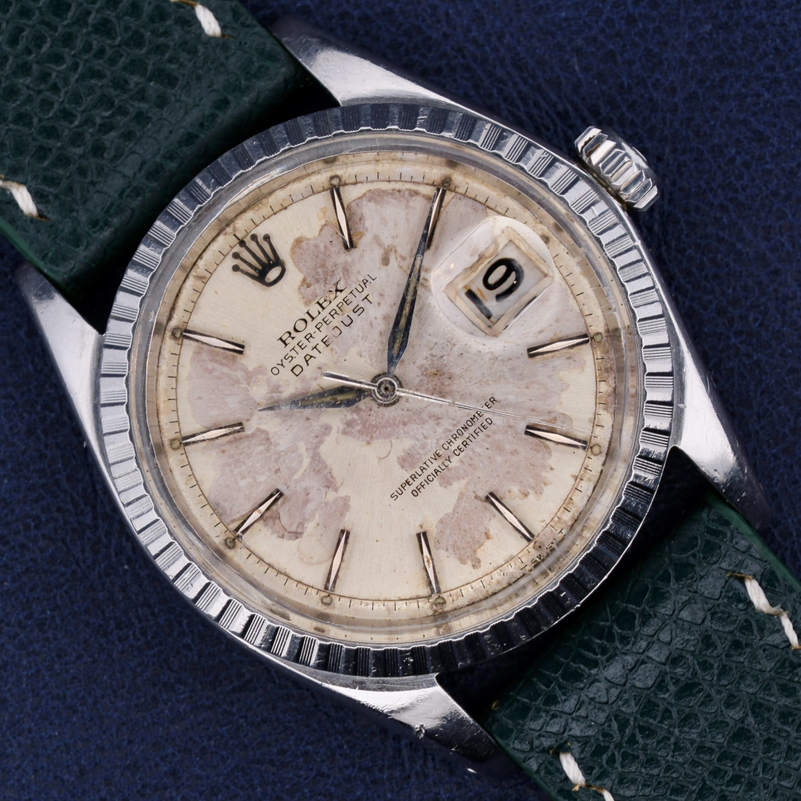 The Rolex Datejust 1603 Pie-Pan Dial features a silver bezel, green leather strap, weathered dial, and date display at 3 oclock, with black hour markers and hands on a blue textured background.