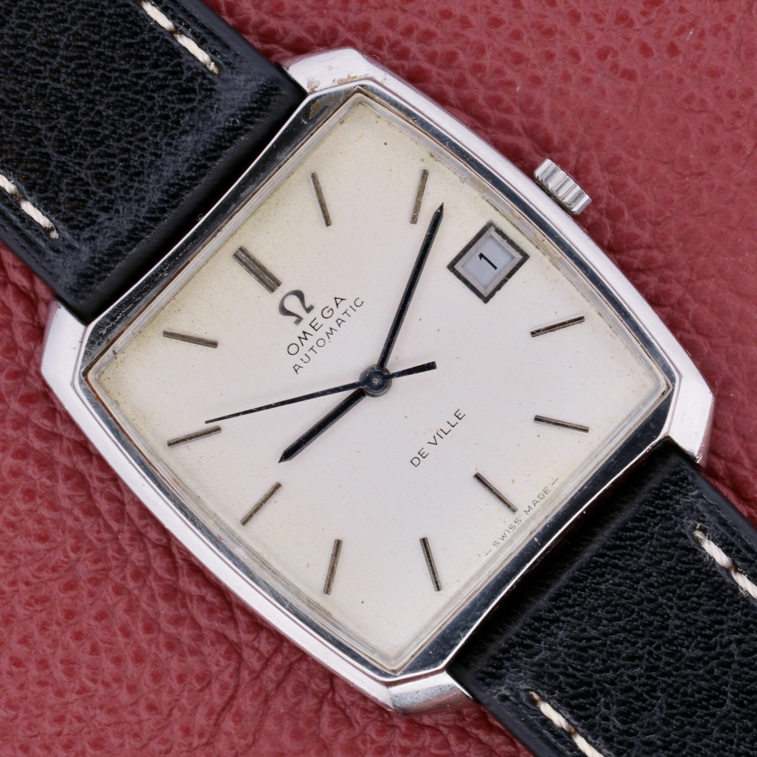 The Omega Deville Automatic Tonneau Case is a vintage watch with a square silver case, cream dial, black hands, date display at 3 oclock, black hour markers, and a black leather strap, elegantly set against a textured red background.