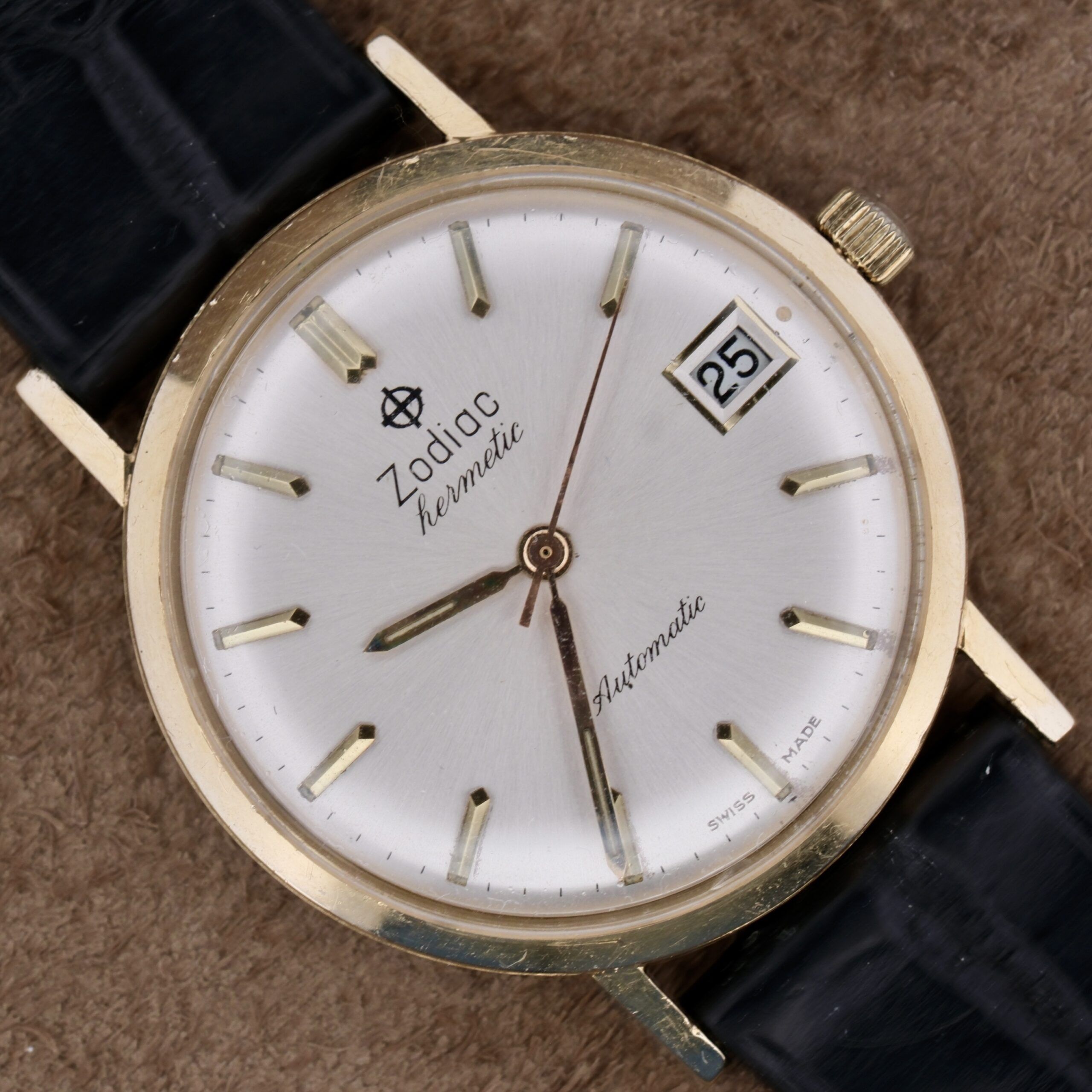 Close-up of a Zodiac Hermetic 14k Gold Automatic Date wristwatch with a gold casing and black strap. The silver face has gold hour markers, shows the date as the 25th, and Swiss Made is inscribed at the bottom.