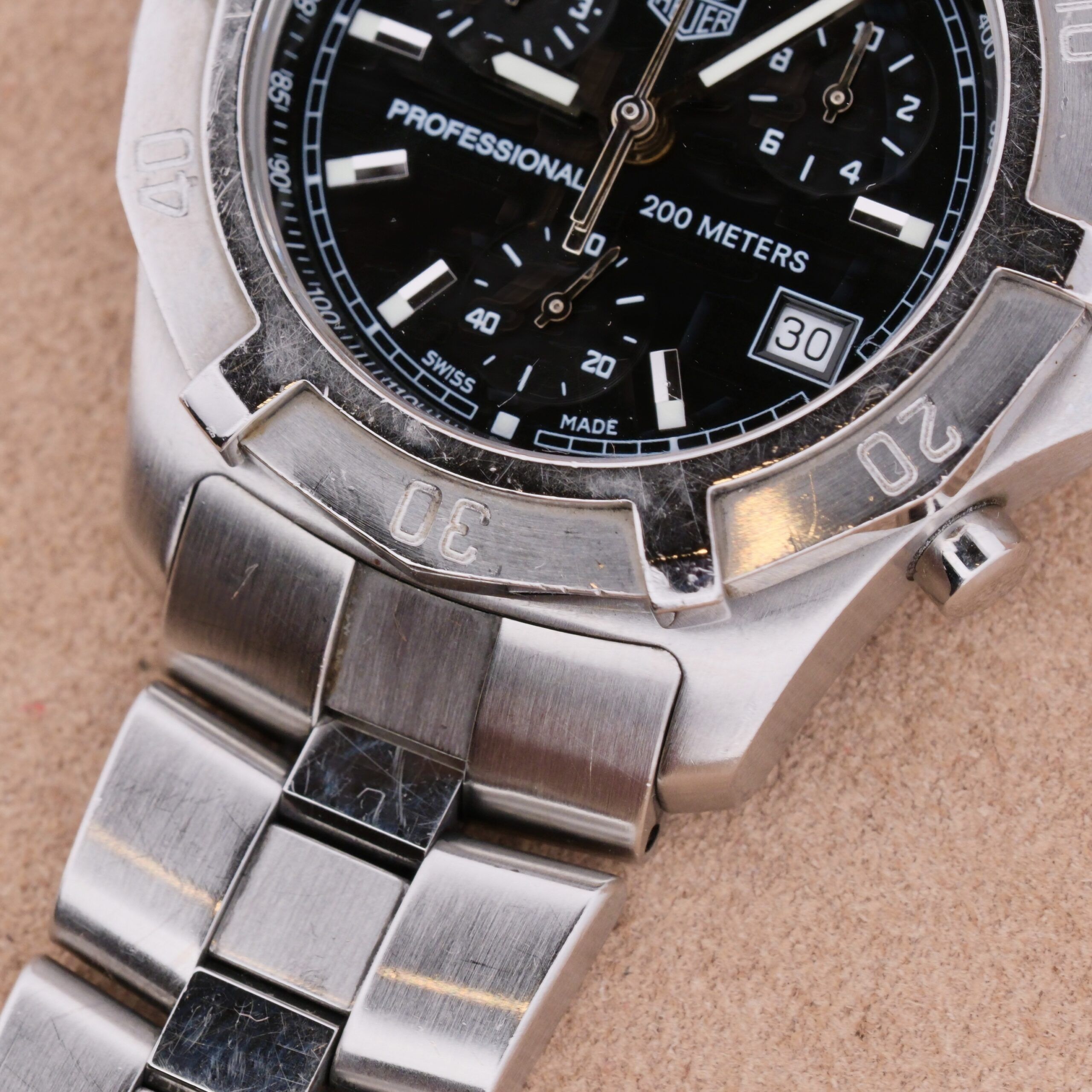 Close-up image of a Tag Heuer 2000 Professional Chronograph: silver watch, black dial, date and time display, 200m water resistance. It features a stainless steel band and unique bezel markings with the brand partially visible.