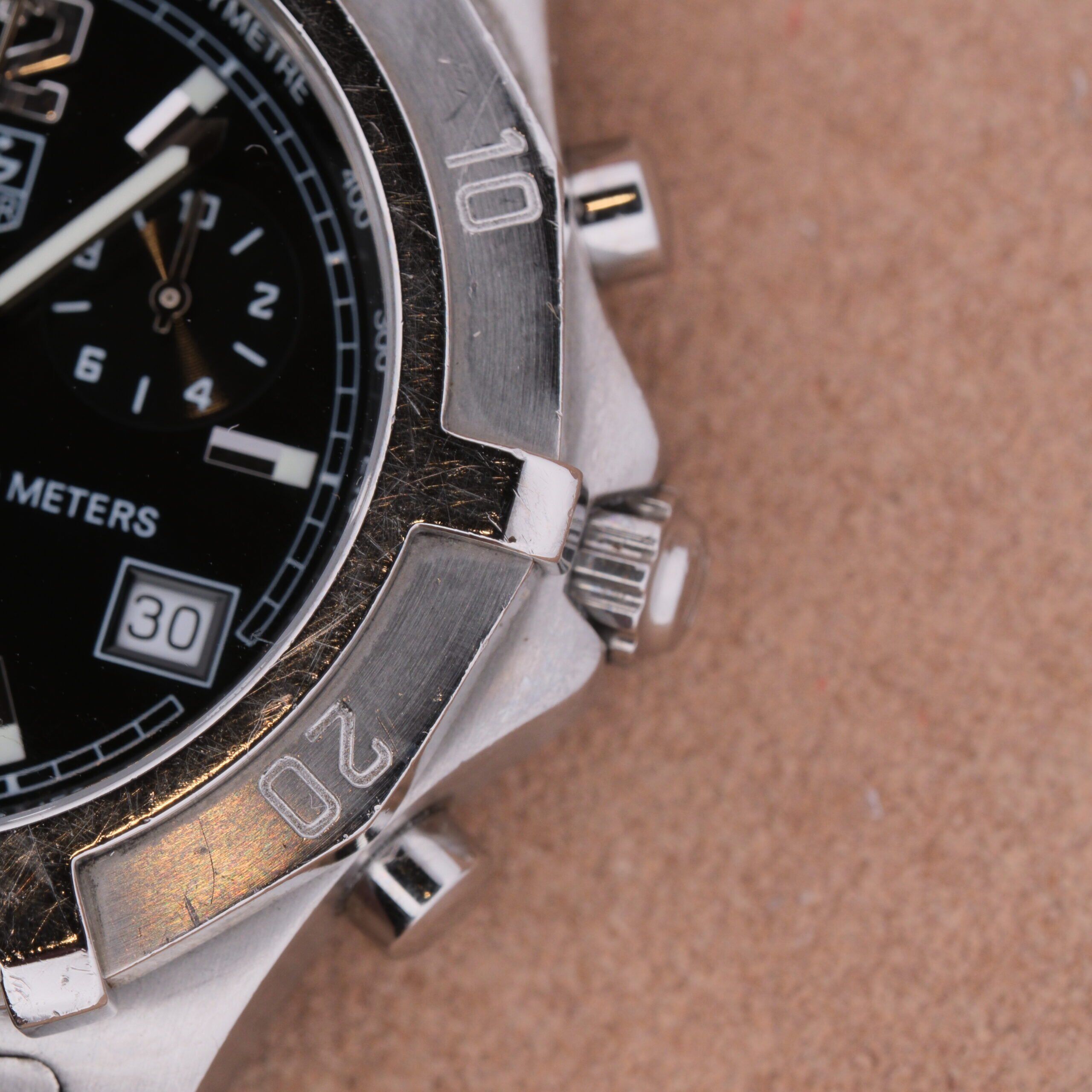 Close-up of a Tag Heuer 2000 Professional Chronograph silver watch with a black dial, featuring three subdials, a date window showing 30, metal markers on the bezel, and protruding buttons. It rests against a textured background, highlighting its sophistication.
