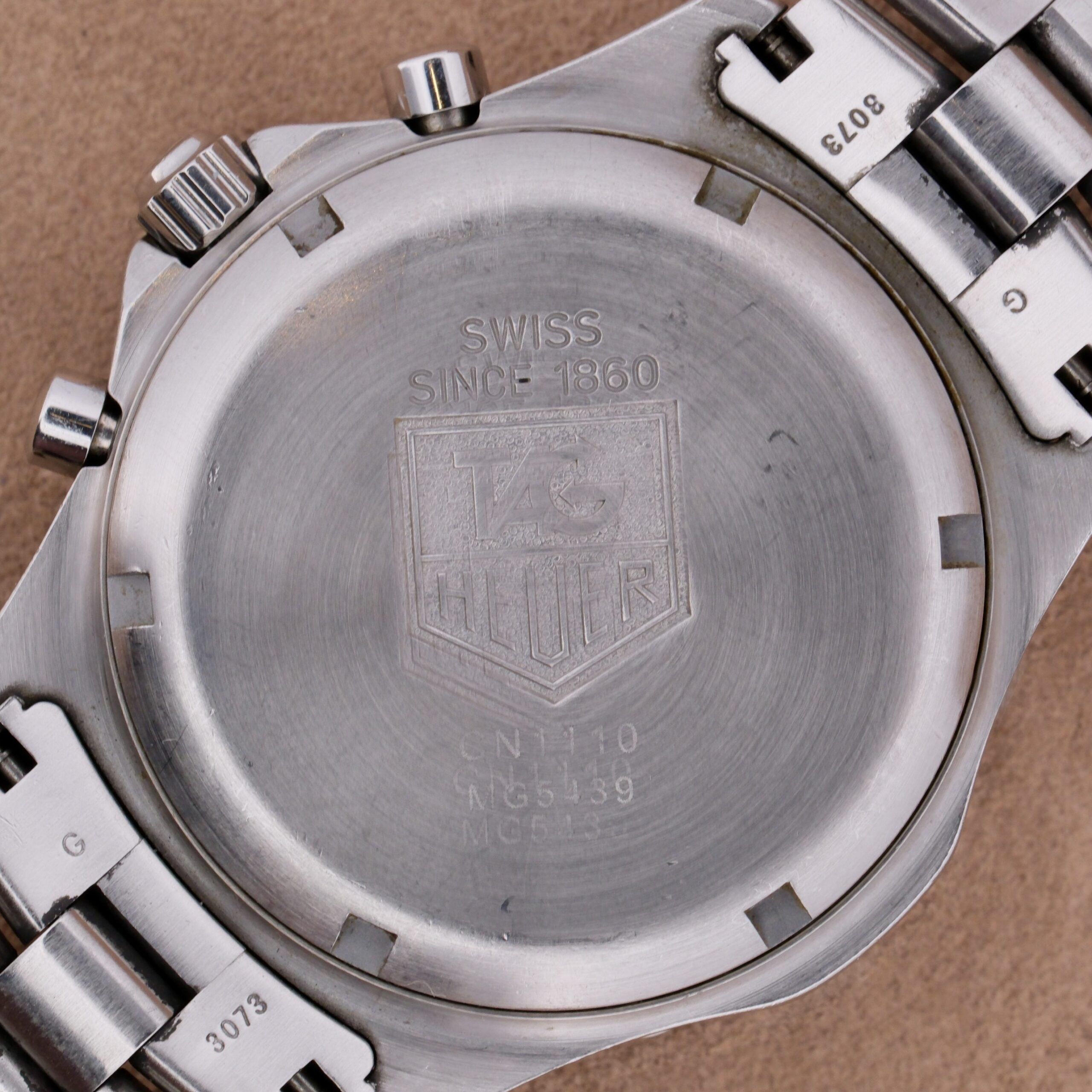 A close-up reveals the back of a Tag Heuer 2000 Professional Chronograph. The metal back is engraved with SWISS SINCE 1860 and the TAG Heuer logo. It features a stainless steel bracelet with visible serial and reference numbers.