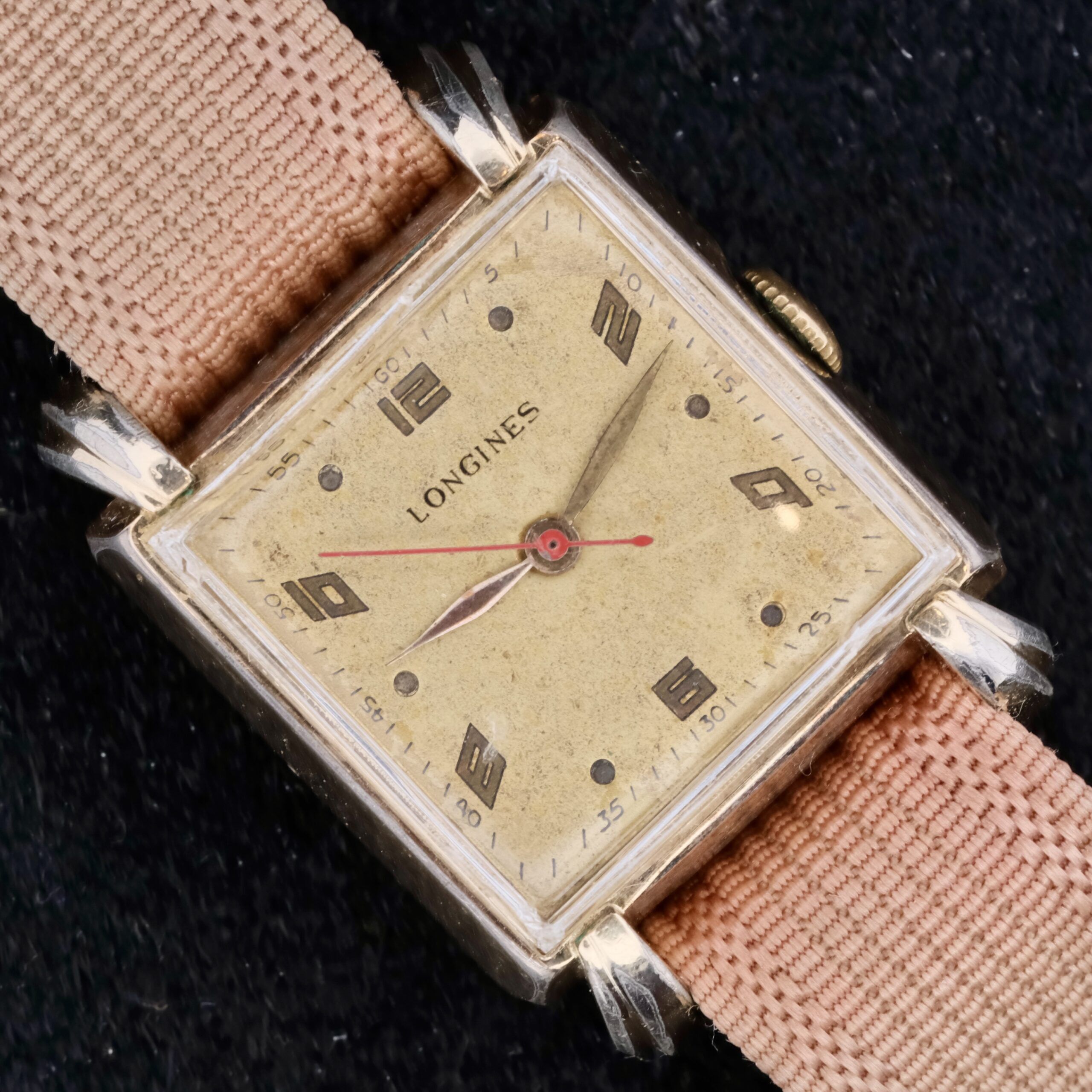 The Longines Square Gilt Dial Central Red Second watch features a gilt dial with Arabic numerals and slim hands. It includes a pinkish-beige fabric strap and gold-toned case, set against a dark textured background.