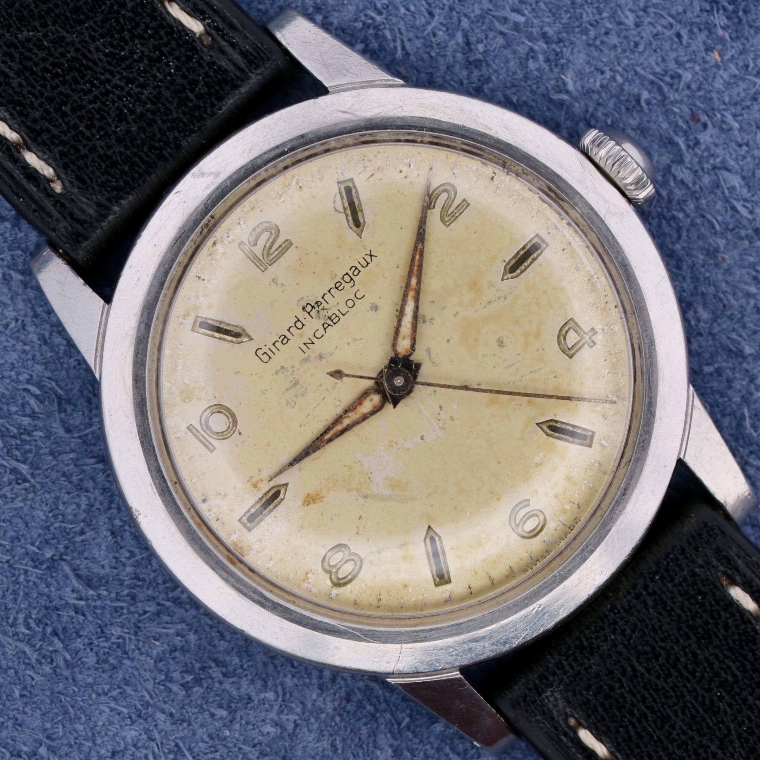 The Girard Perregaux Incabloc Manual Wind wristwatch features a beige face with silver hour markers and hands at approximately 10:10, a black leather strap, and visible signs of aging on the face. The brand name is clearly displayed.