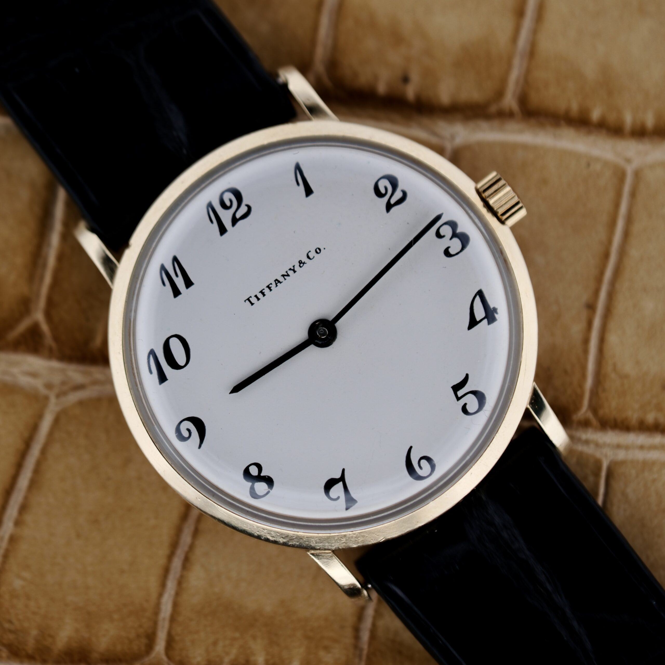A close-up of the Tiffany & Co. Arabic Numerals Dial T-101 showcases its elegance with a gold case and black leather strap. The watch features a white dial with black numerals and sleek hands on a textured beige backdrop, highlighting its refined craftsmanship.