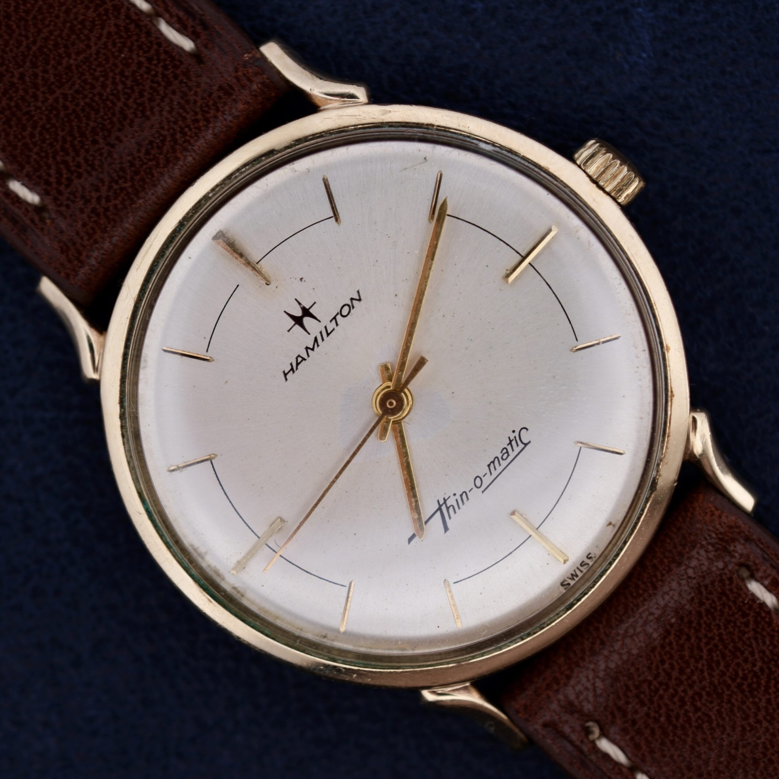 The Hamilton Thin-O-Matic Sunburst wristwatch features a gold case, sunburst silver dial with gold hour markers and hands, a brown leather strap, and Swiss branding beneath the hands on a blue background.
