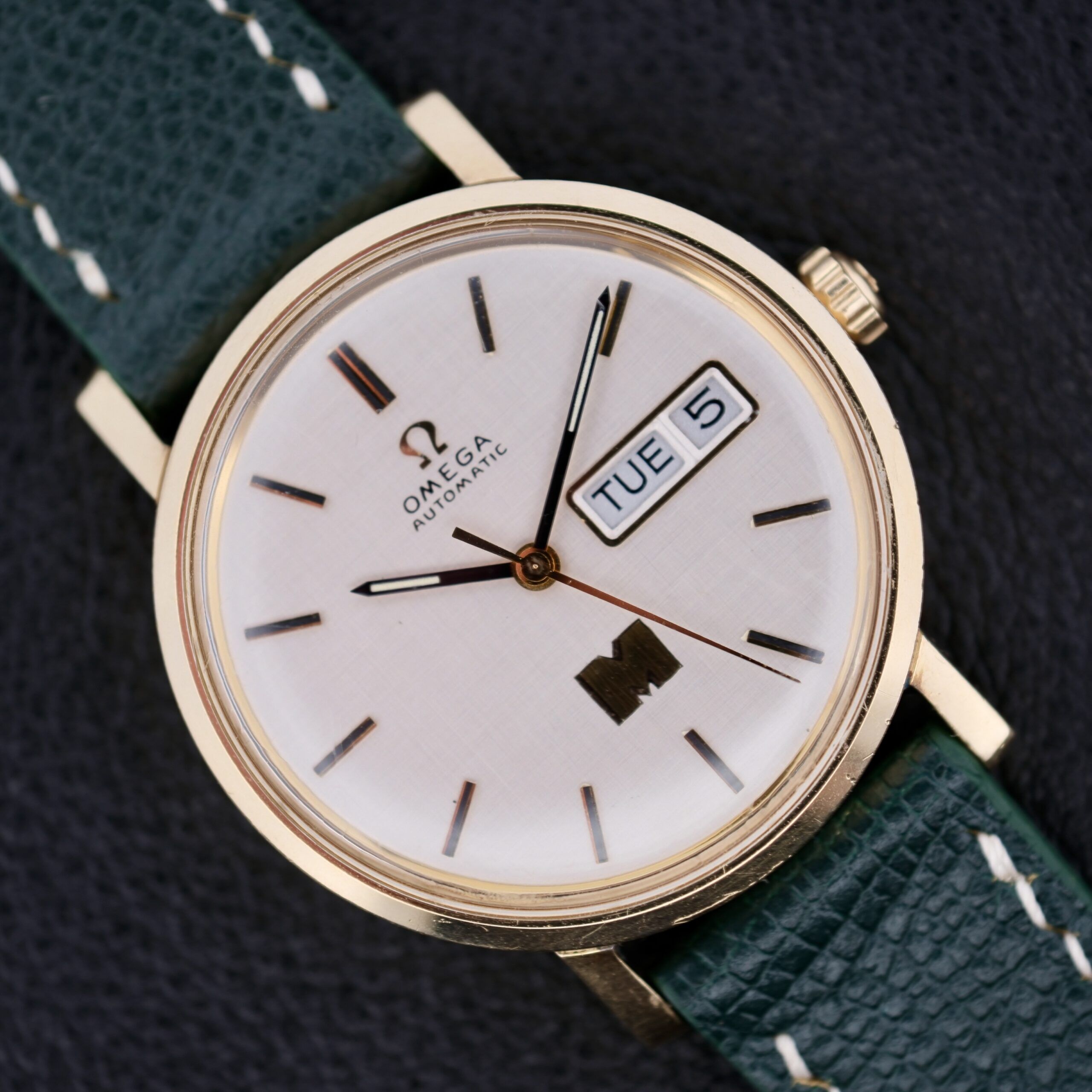 A close-up of the Omega Automatic Day/Date Linen M Dial watch, featuring a gold case and green leather strap. The elegant Linen M Dial displays the time, Tuesday, and 5th against a textured dark background.