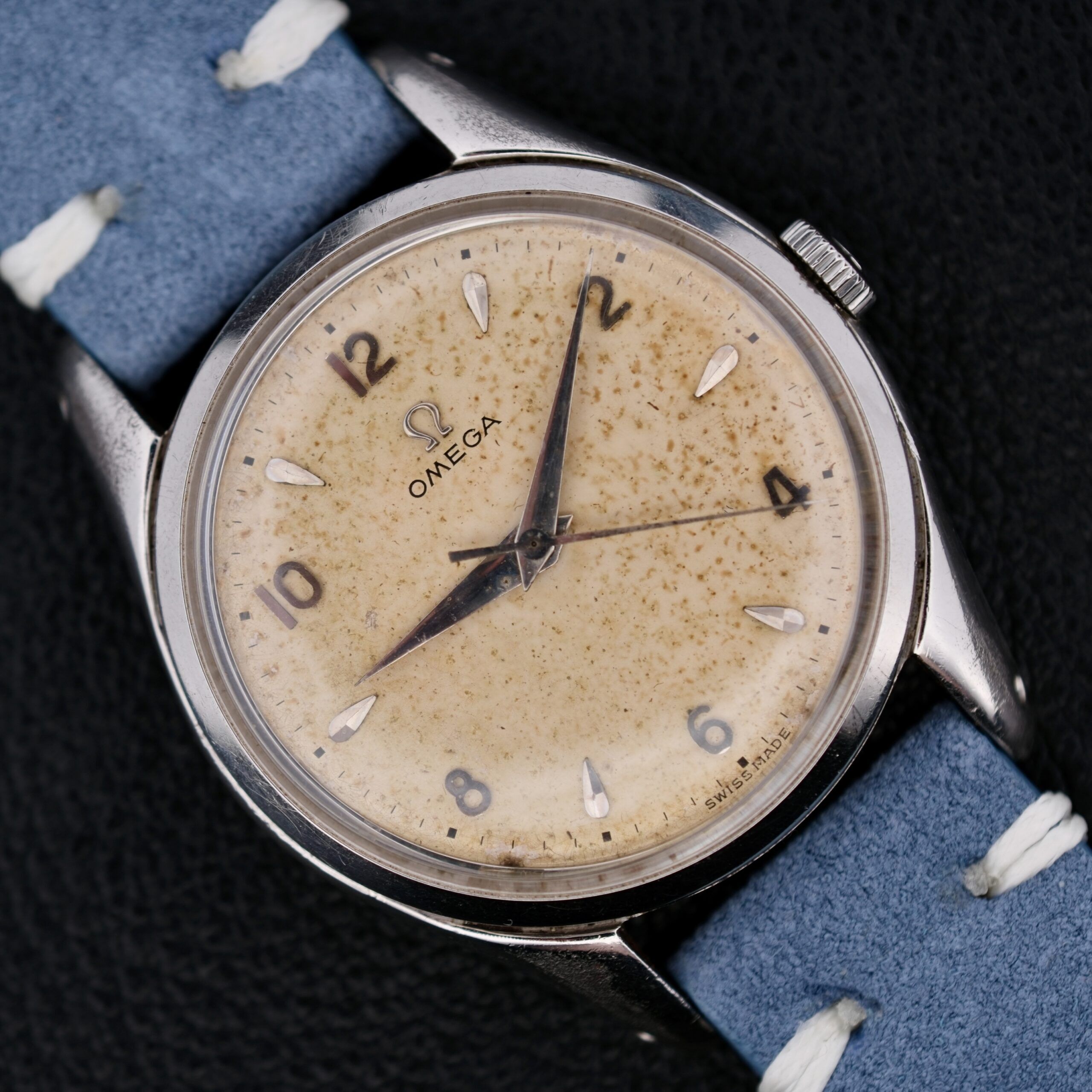 Close-up of an Omega Pre Seamaster Cal. 283 wristwatch with a weathered cream dial, dark hour markers, and two black hands, set in a metallic case with a light blue textured strap against a dark background.