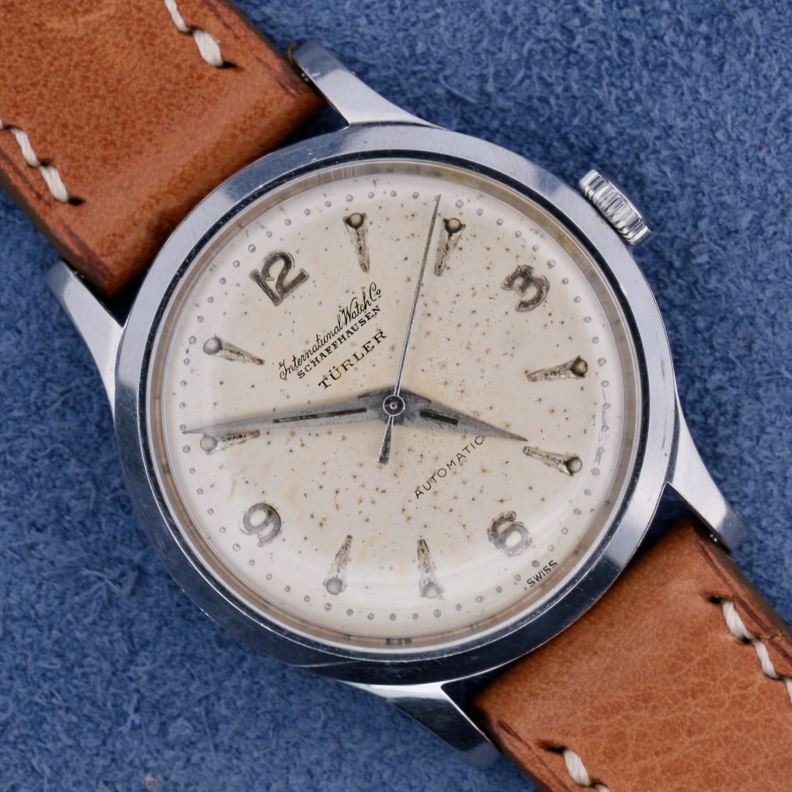 The IWC Automatic Turler Dial C. 852 is a vintage wristwatch featuring a speckled cream Turler dial with Arabic numerals and silver hands. It has a stainless steel case, tan leather strap, and the brand name Automatic is elegantly displayed on the dial against a blue fabric backdrop.
