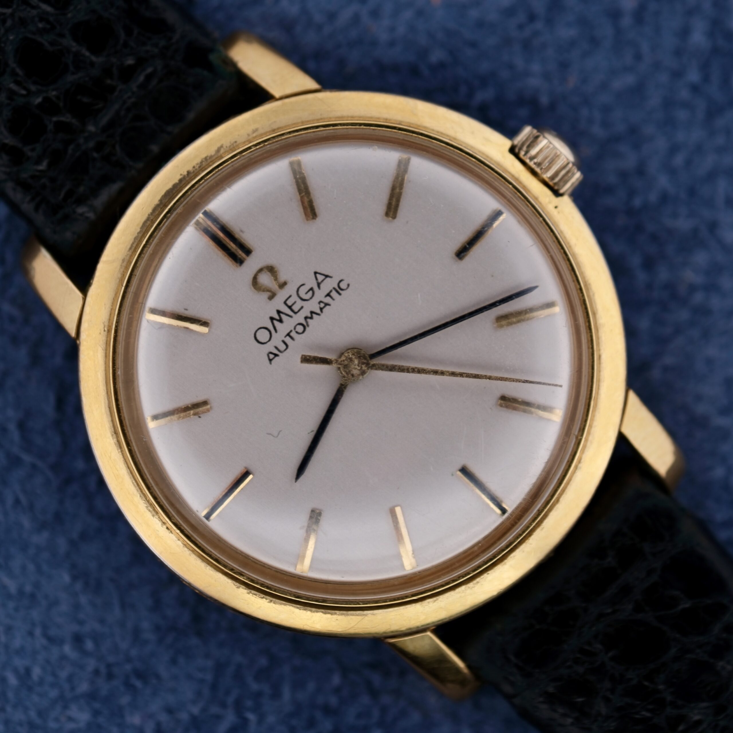 A close-up of the Omega Automatic Ladies wristwatch showcases its gold case, white dial, and black leather strap. It features elegant gold hour markers and hands on a striking blue textured background.