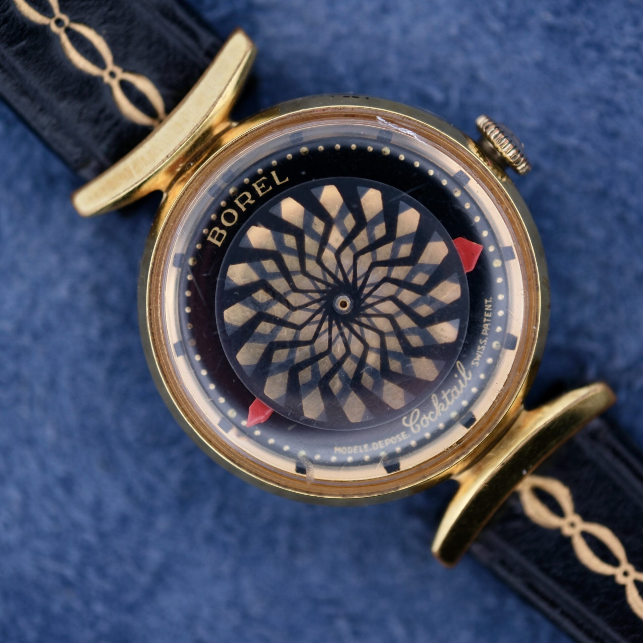 An Ernest Borel Kaleidoscope Cocktail watch with a black and gold spiral-design dial, red pointers, and a gold bezel. Its black leather strap is intricately patterned and elegantly placed on a blue textured surface.