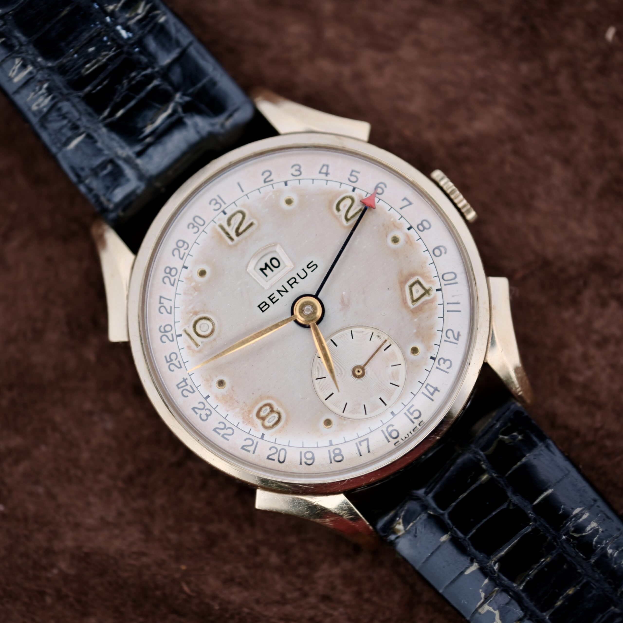 The vintage Benrus Day/Date Pointer watch features a gold-tone case and black crocodile-patterned leather strap. Its face has gold numbers, a small second subdial, and a date pointer set to the 10th against a brown fabric background, enhancing its timeless elegance.