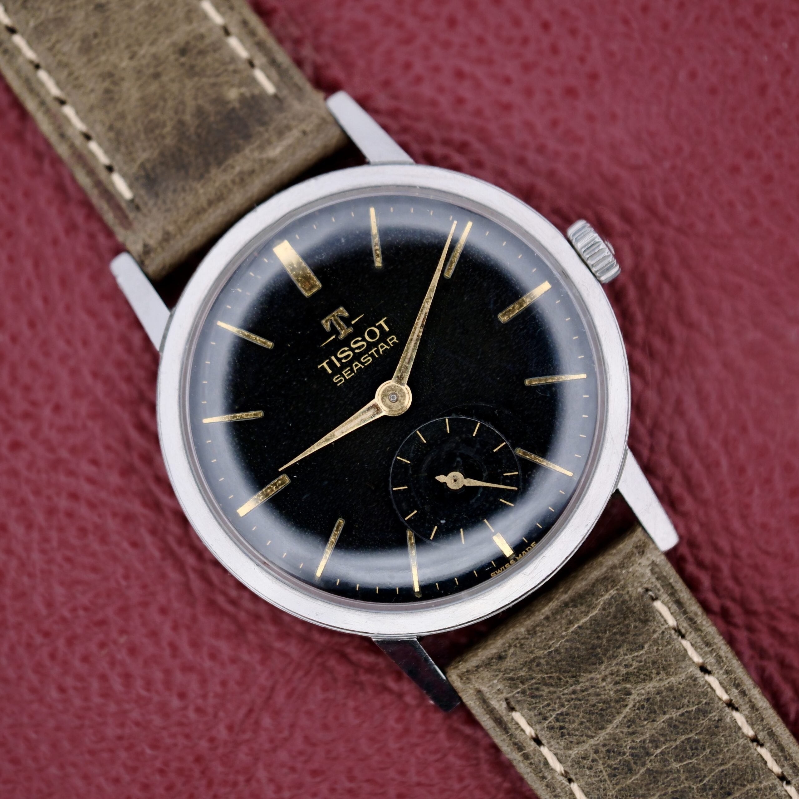 The Tissot Seastar Black Gilt Dial Sub Second is displayed with its black dial, gold hour markers, small second subdial, and leather strap on a textured red surface.