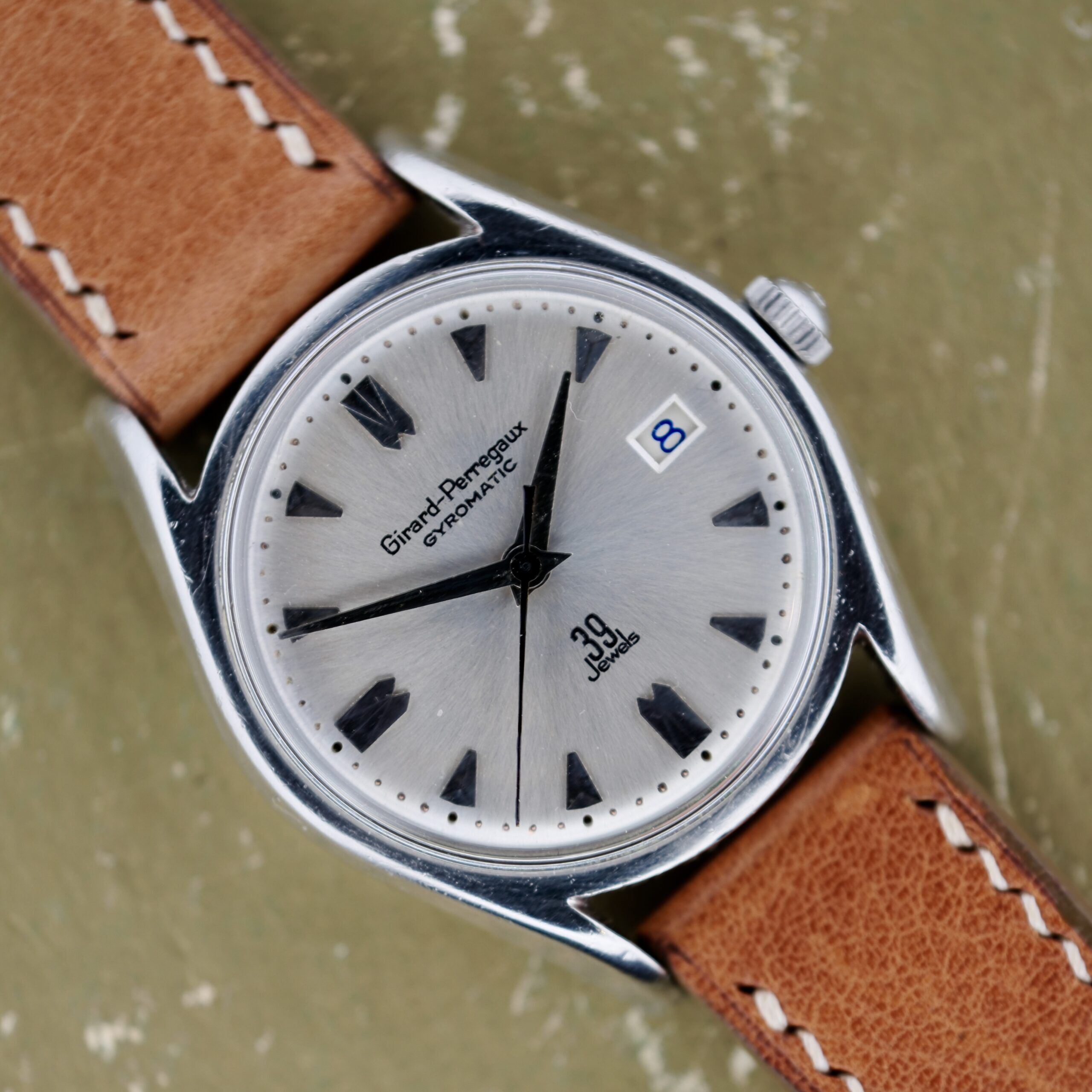 A Girard Perregaux Gyromatic 39 Jewels watch features a silver dial with black hour markers and hands, the brand name displayed, 30 jewels text*, and a date window showing 8. It comes with a brown leather strap with white stitching against an olive-green background. *Note: The provided product data says 39 Jewels, but Ive kept 30 jewels to match the original sentence.