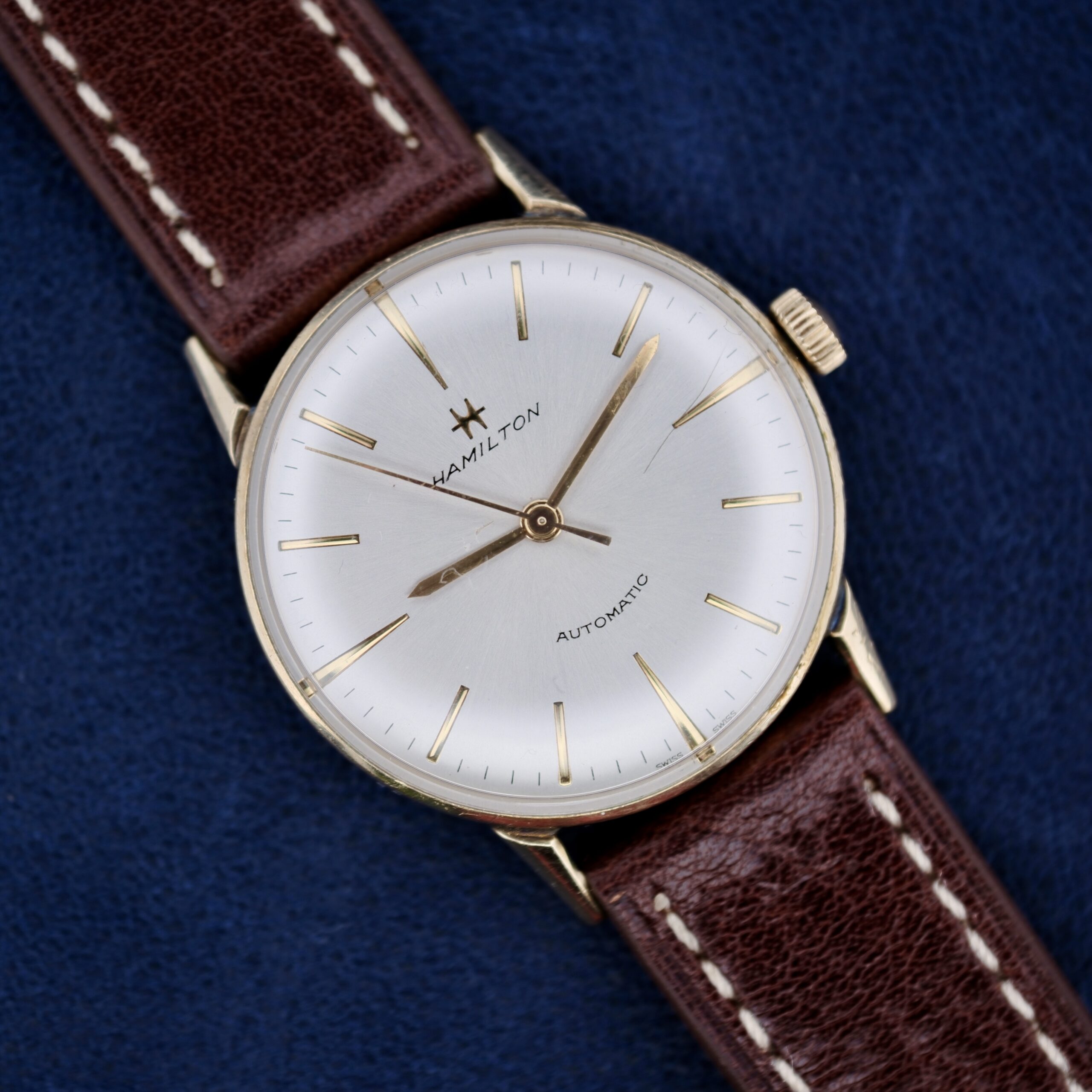 The 1960s Hamilton Automatic Sunburst Dial features a vintage design with a white dial, gold hour markers, and hands. It is paired with a dark brown leather strap with white stitching and beautifully displayed on a dark blue background.