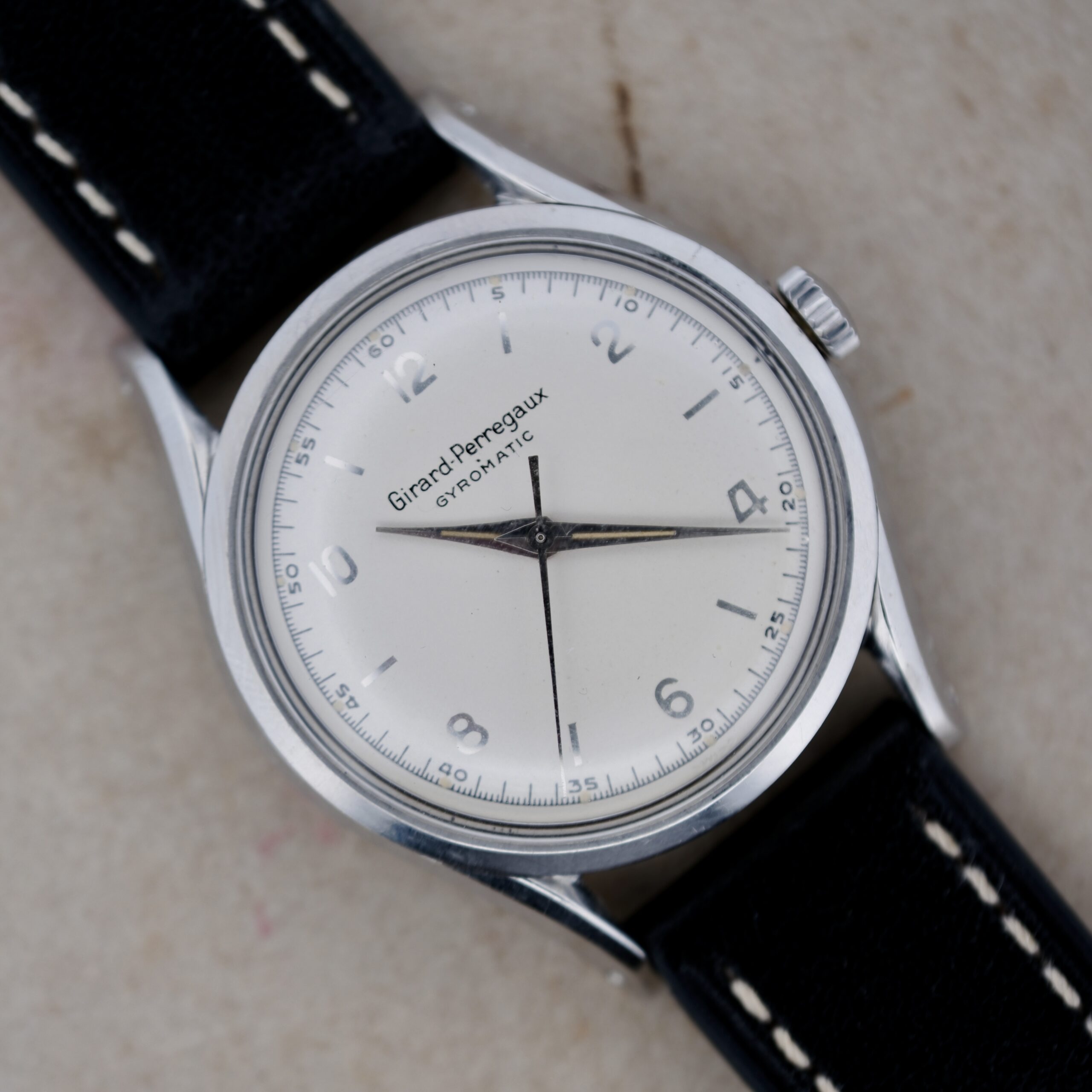 The vintage Girard Perregaux Gyromatic 6169 Caliber 47 wristwatch boasts a white dial with silver hour markers and elegant black hands. The classic silver case pairs beautifully with a refined black leather strap accented with white stitching.