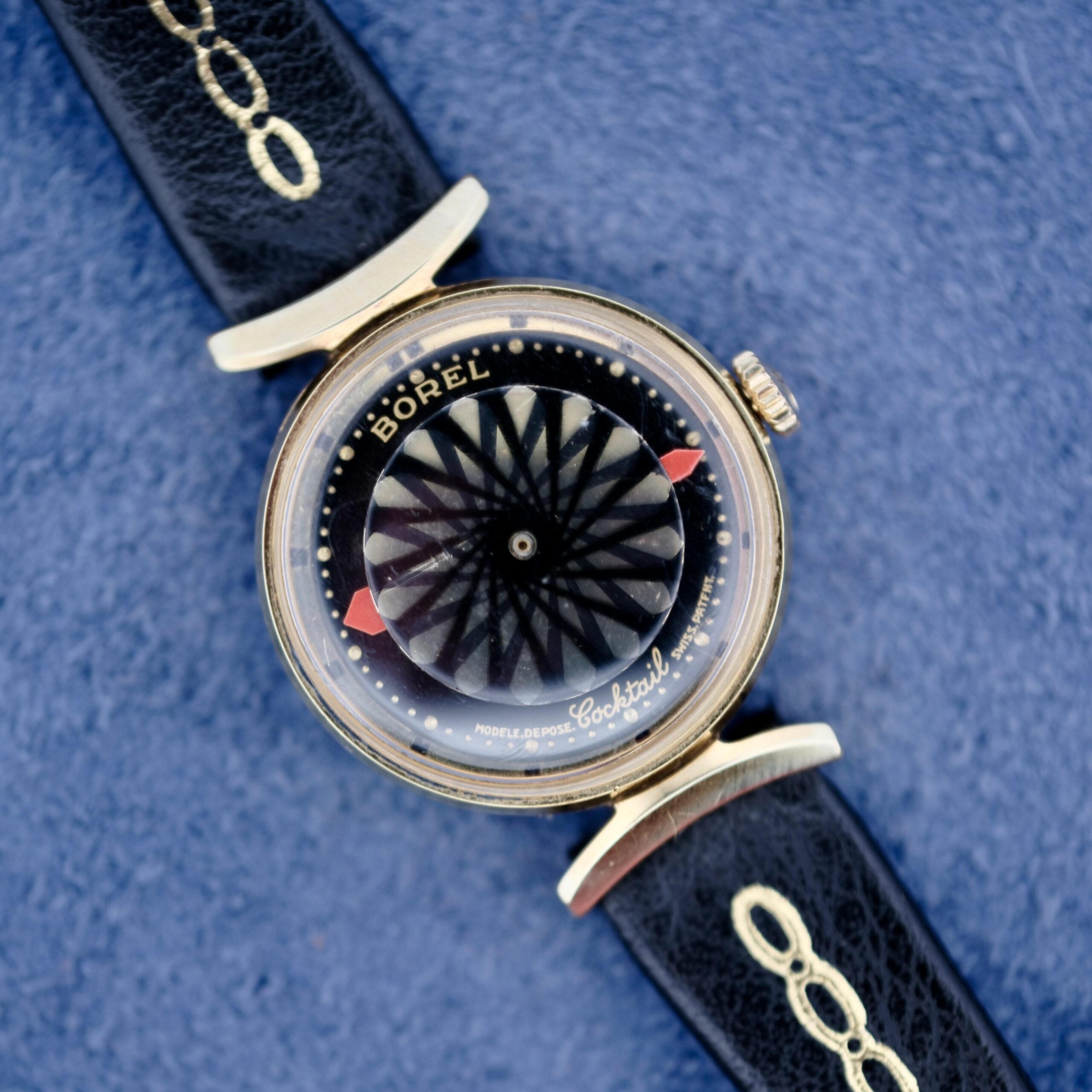 The Ernest Borel Kaleidoscope Cocktail Black Dial watch features a vintage gold-toned round case, a striking black geometric dial with red hour markers, and a textured blue background, paired with a black leather strap accented in gold.