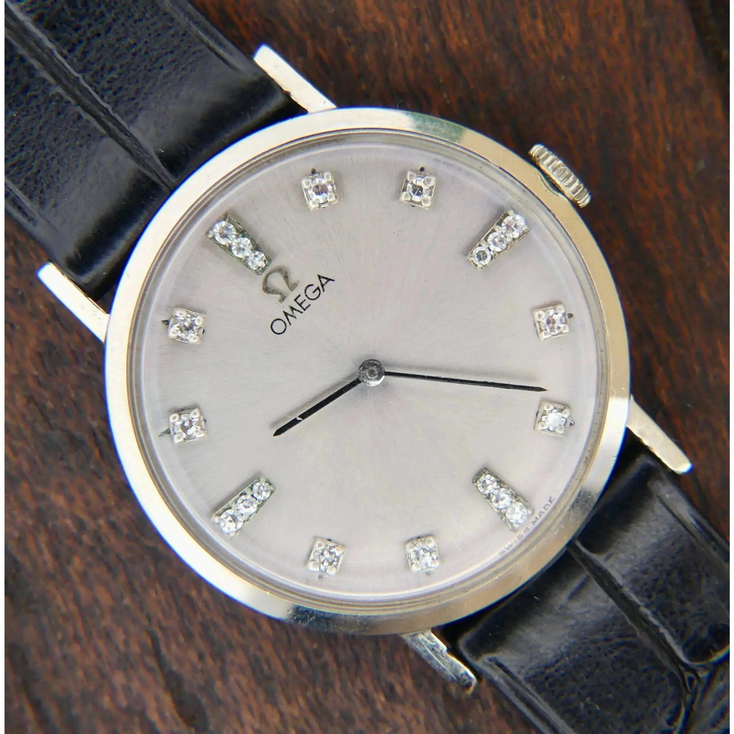 The 1965 Omega Deville 14k Ref. D6672 wristwatch, with silver hour markers and diamond accents, is displayed on a wooden surface. It features a round face, a black leather strap, and two black hands for hours and minutes.