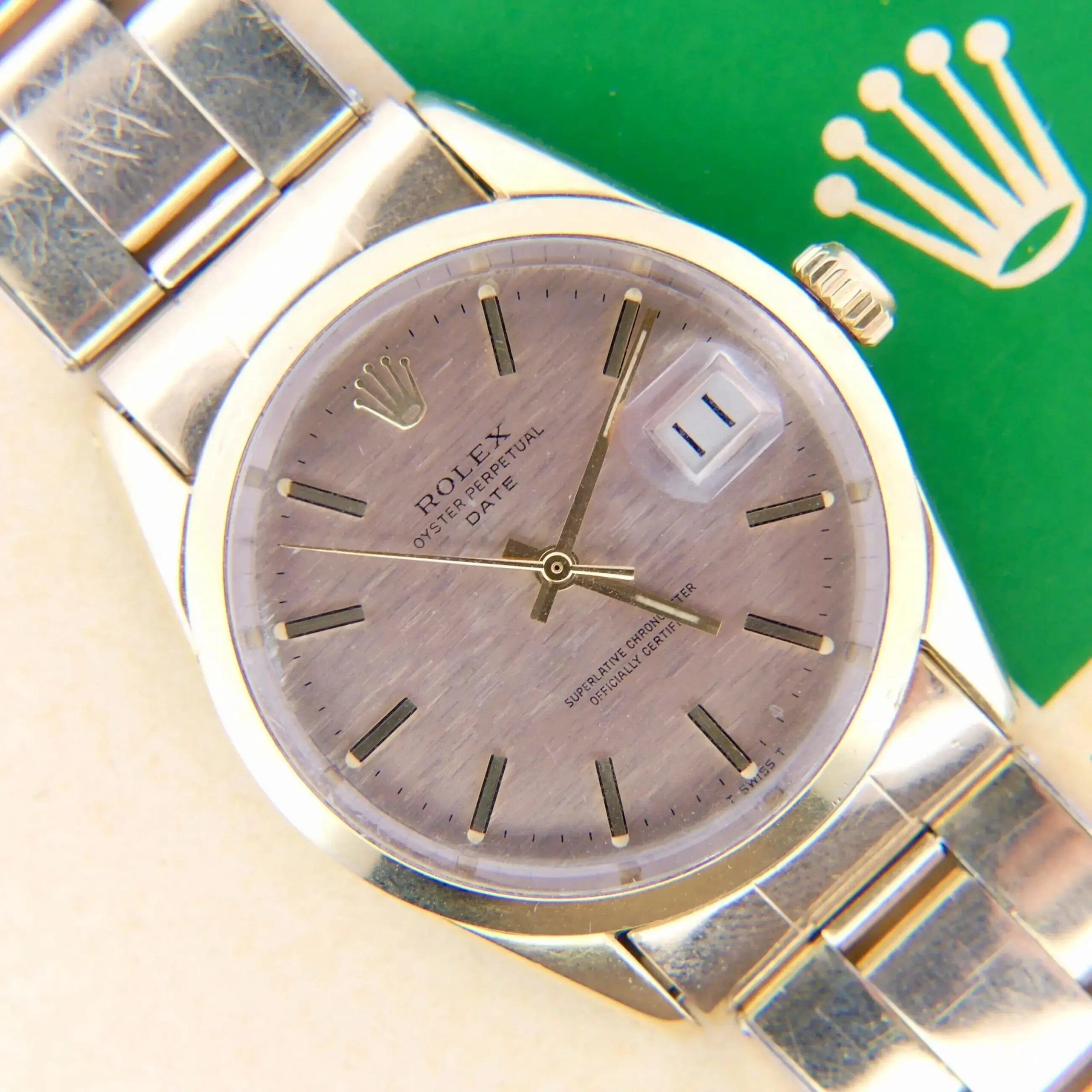 Close-up of a 1978 Rolex 1550 Oyster Perpetual Date watch. It features a brushed silver dial with gold hour markers and a date window at three oclock, along with a green card displaying the crown logo.