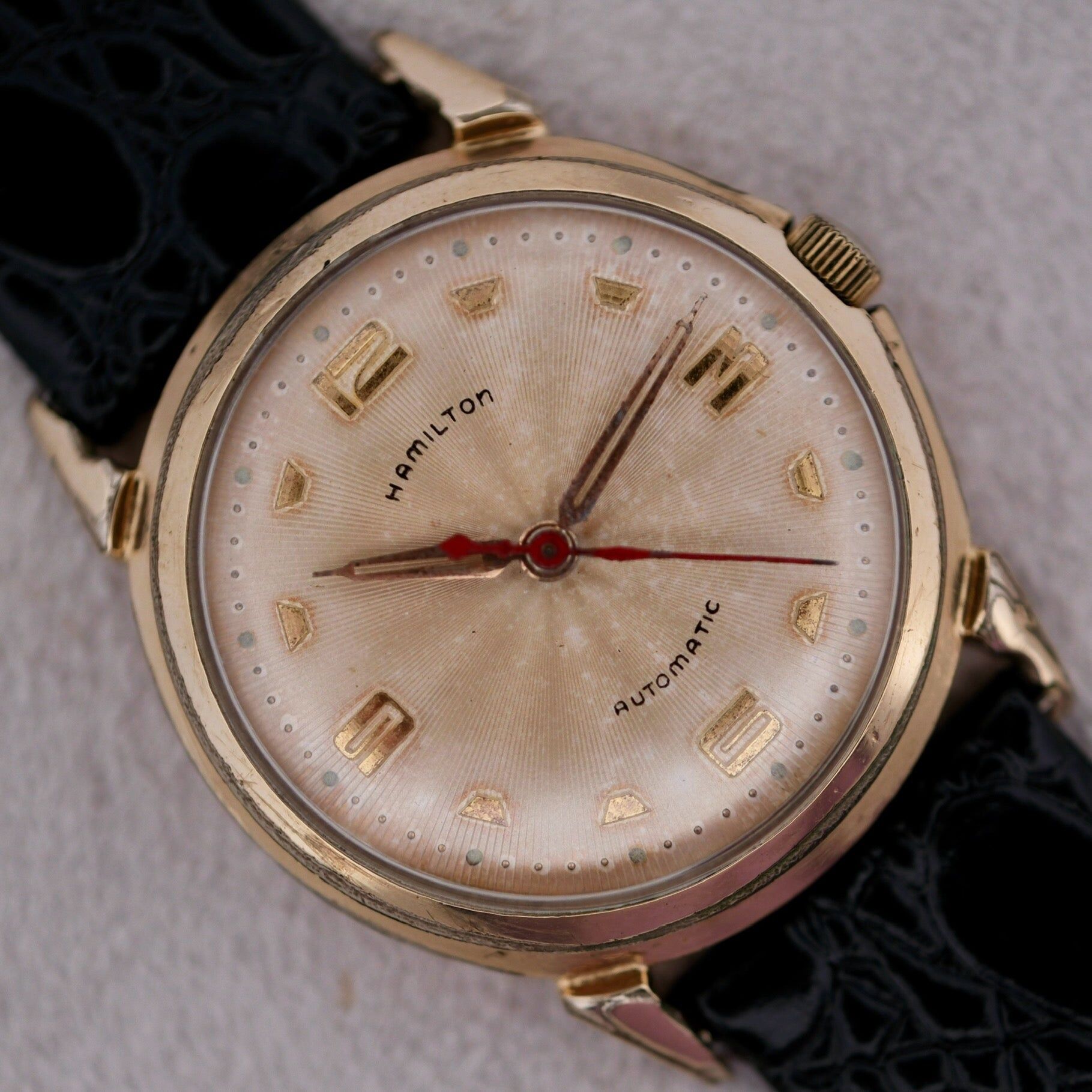 A vintage Hamilton Accumatic III Cal. 672 wristwatch featuring a gold-tone case, black leather strap, off-white dial with gold numerals and hands, a red second hand, and the word Automatic.