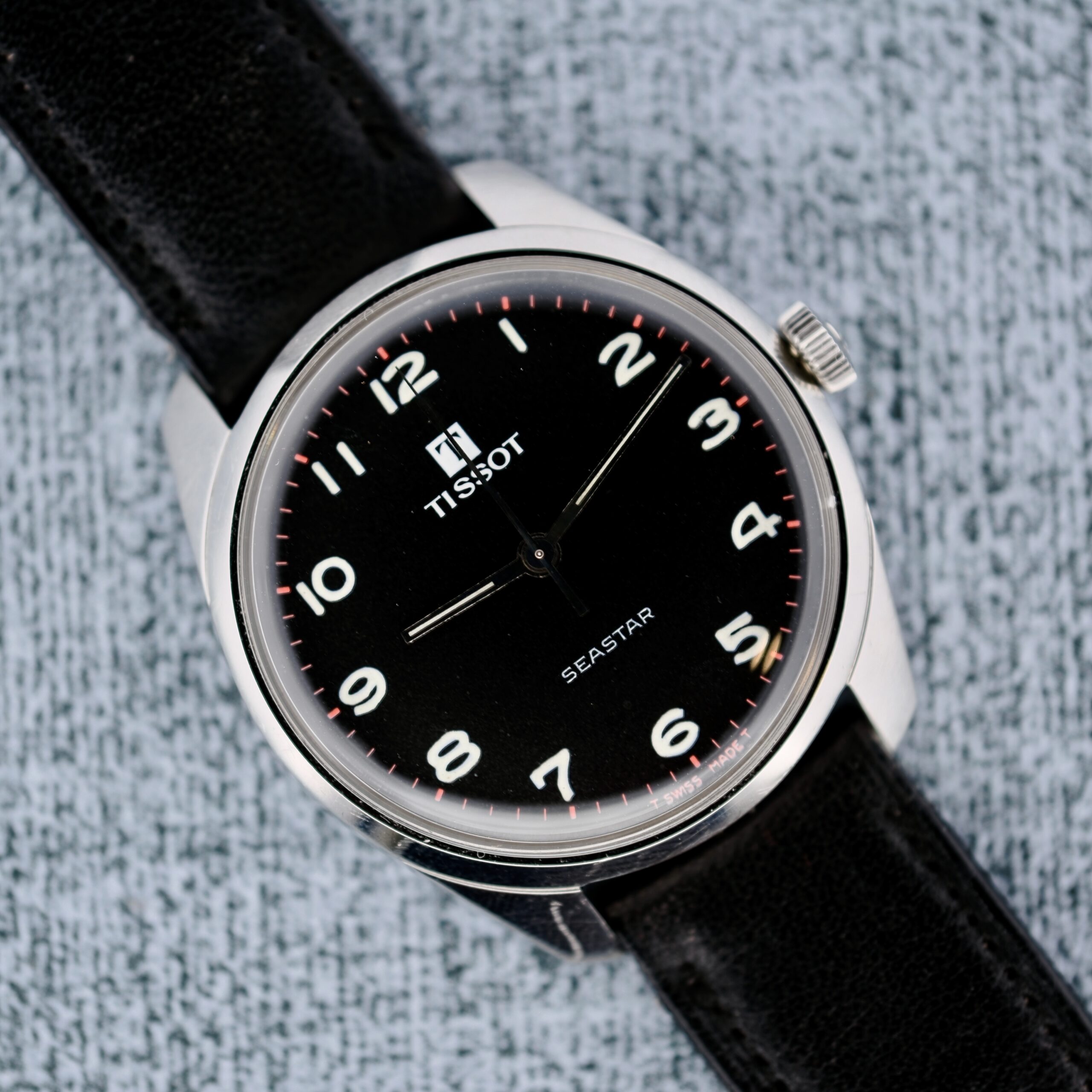 The Tissot Seastar Manual Wind Arabic Numerals features a textured light gray background, black leather strap, and black face with white numerals and hands.