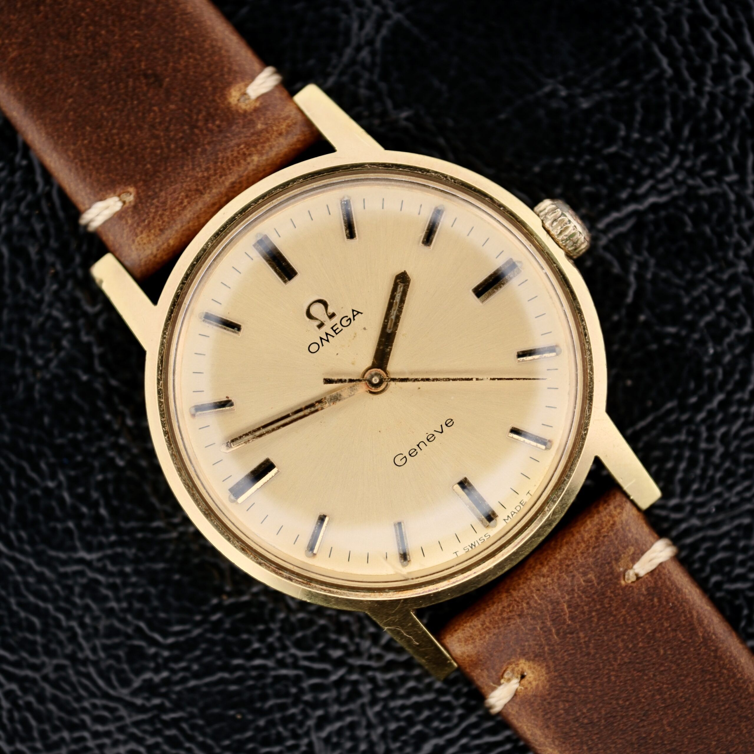 This vintage Omega Geneve Gold Champagne Dial 135.070 features a gold-tone case, brown leather strap, and light-colored dial with black hour markers and hands. The Omega logo and Genève are displayed on the dial against a black textured leather background.