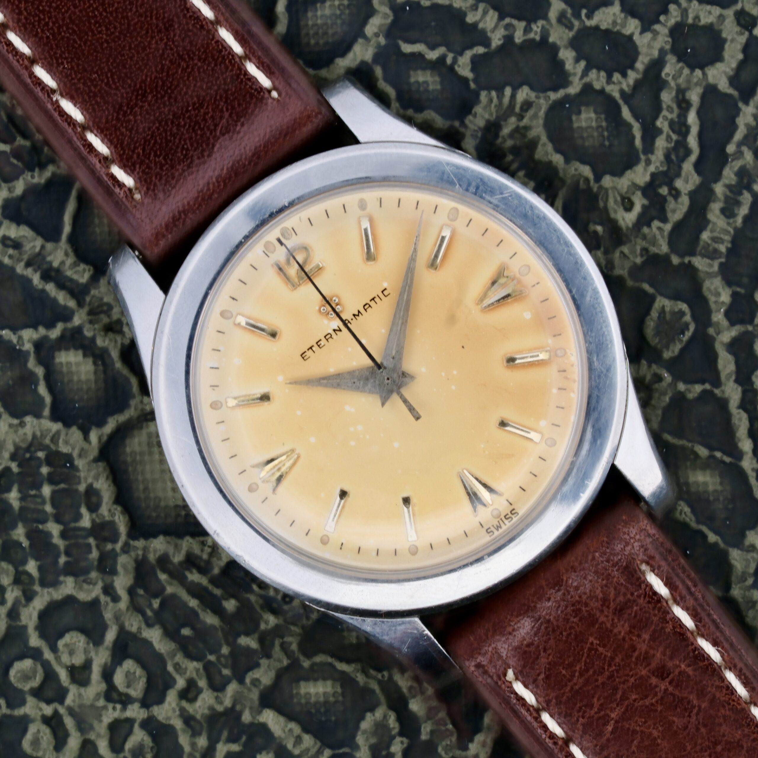 The Eterna-Matic Tropic Dial Hidden Crown is a vintage wristwatch featuring a yellowed dial with Roman numerals, silver casing, and brown leather straps with white stitching. The brand name Eterna-Matic is on the dial against a patterned background.