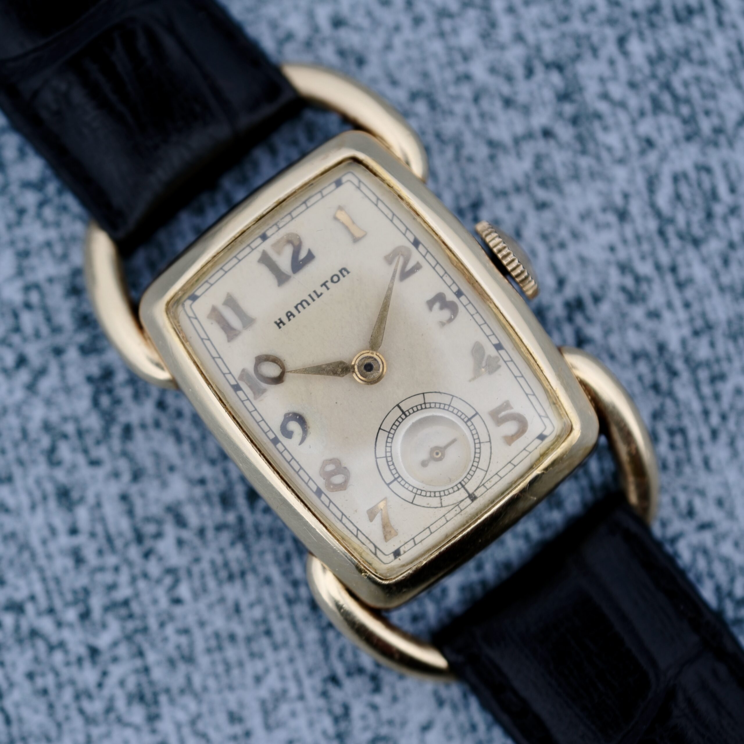 The 1940s Hamilton Bowman wristwatch is displayed on a textured gray surface, showcasing a rectangular gold casing. It features an off-white dial with Arabic numerals, a small subdial, and comes with a black leather strap.