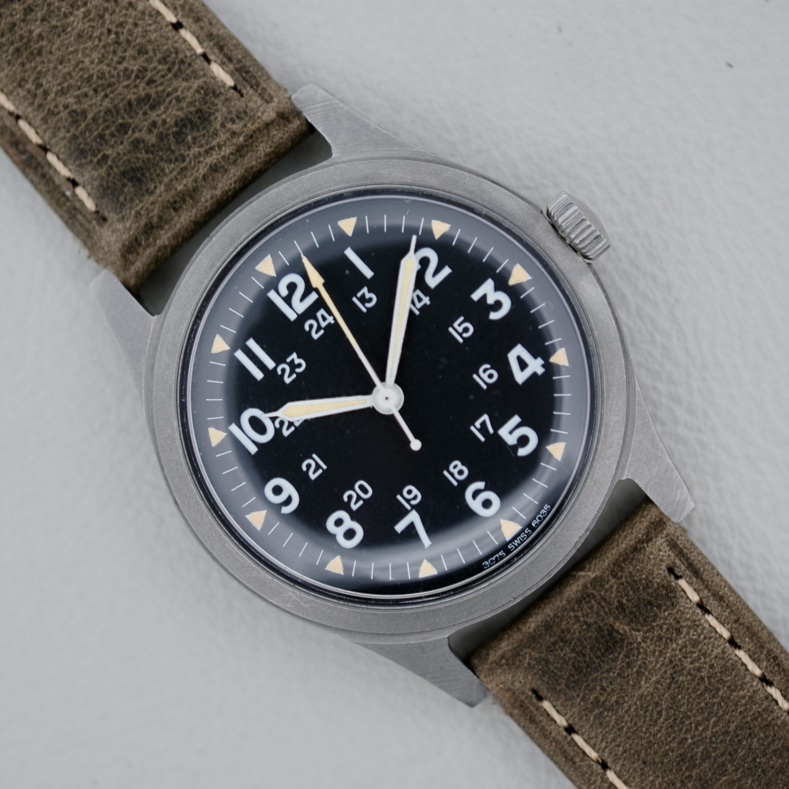 The 1974 Benrus GG-W-113 U.S. Military Issued wristwatch features a black dial with white numerals and hands, including a second hand, brown leather strap with visible stitching, and a brushed metal case against a light gray background.