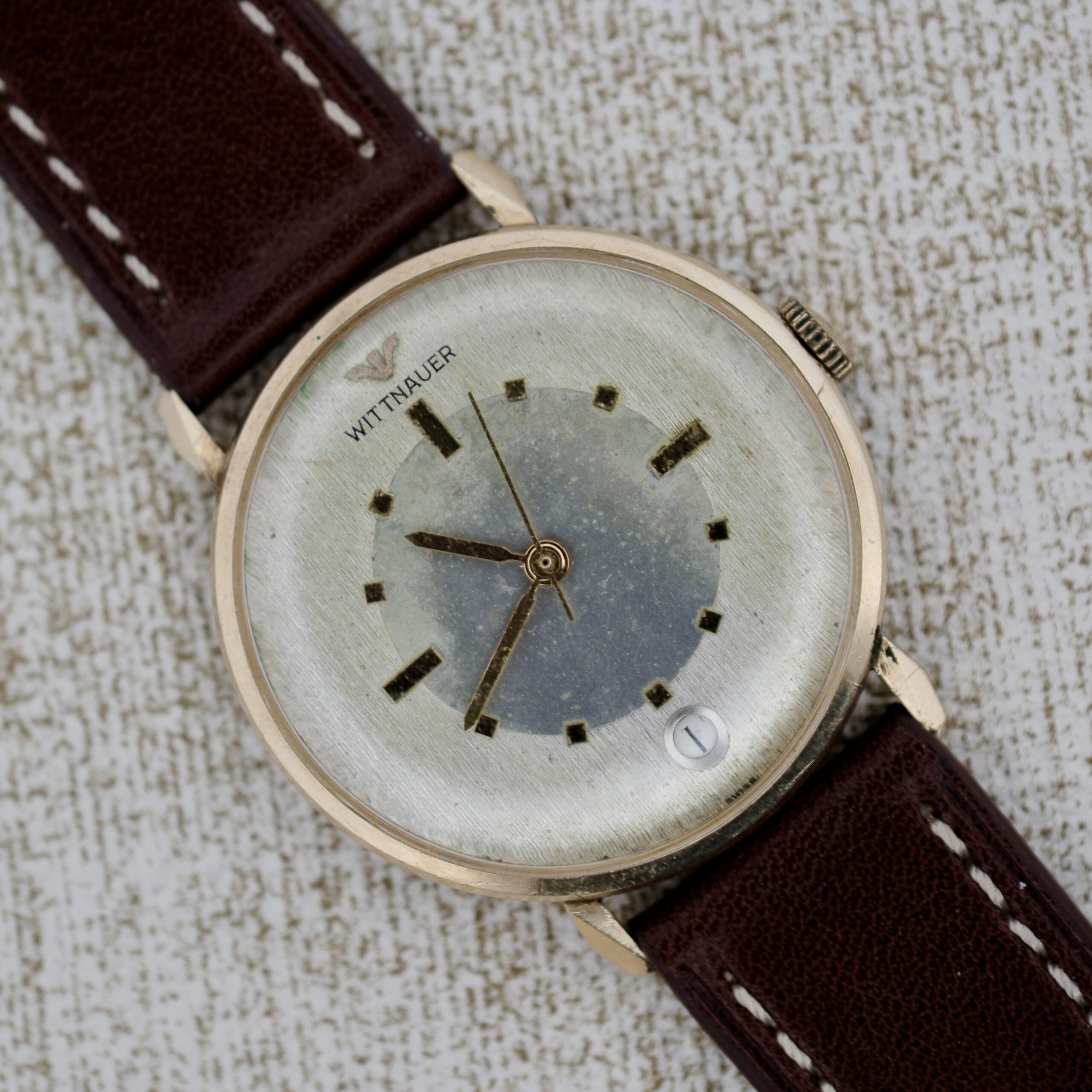 The Wittnauer 627-1-215 Manual Wind Date watch features a round silver-tone dial, gold case, brown leather strap, simple hour markers, date window at 4 o’clock, and the Wittnauer logo below 12 o’clock.