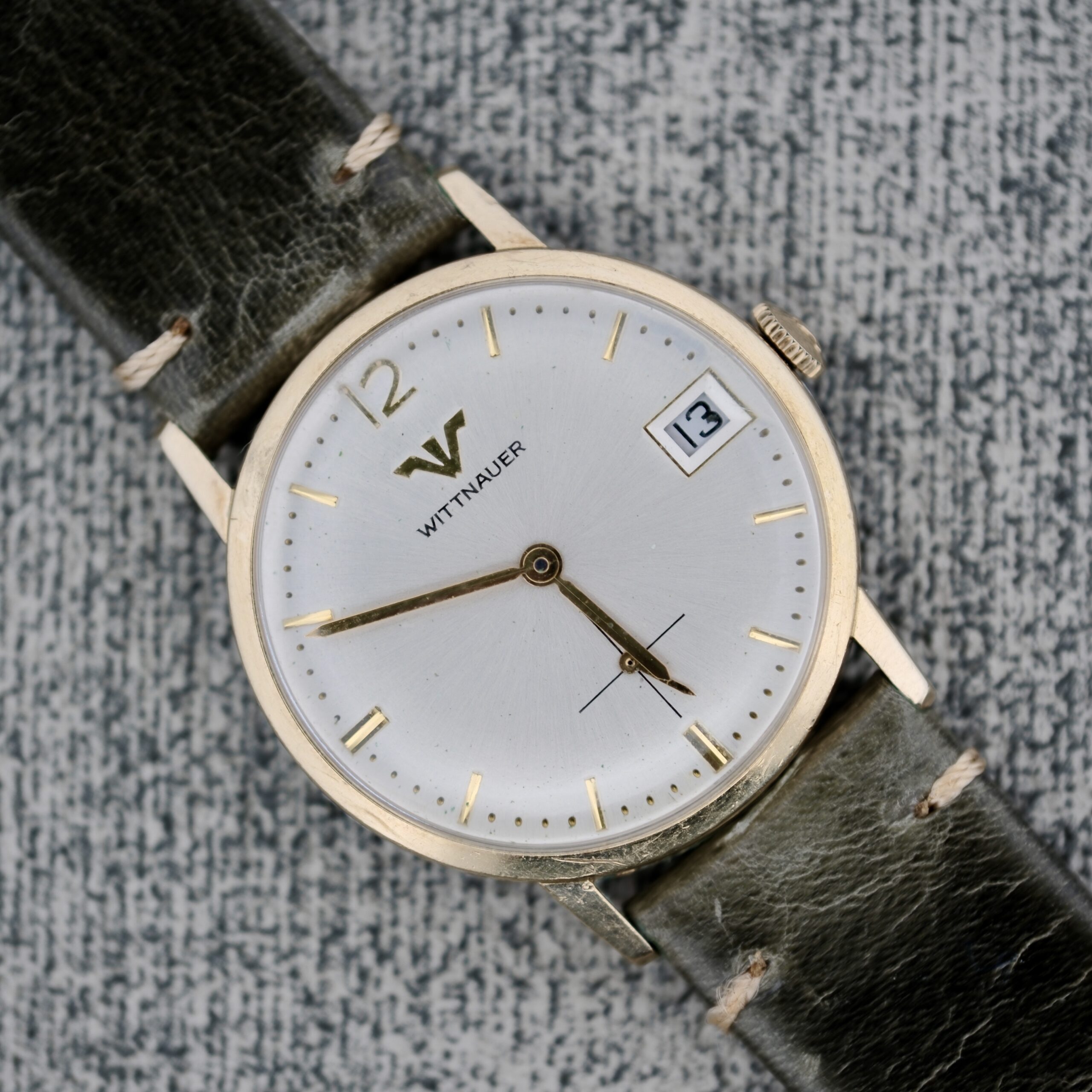 A Wittnauer Date Manual Wind 2732-1-C189 wristwatch with a round silver dial, gold hour markers, date window at 3 o’clock, and a dark green leather strap, shown on a textured gray background.